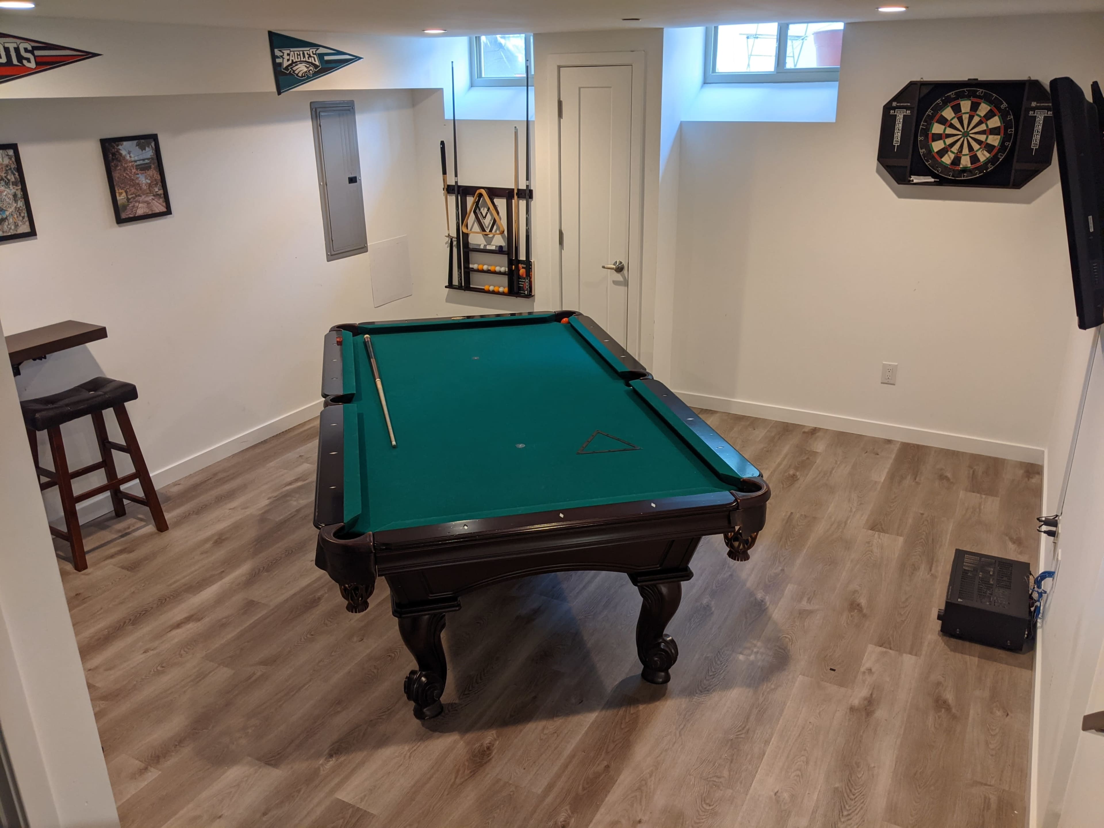 A well-lit basement with a green felt pool table, a dartboard on the wall, and bar stools nearby.
