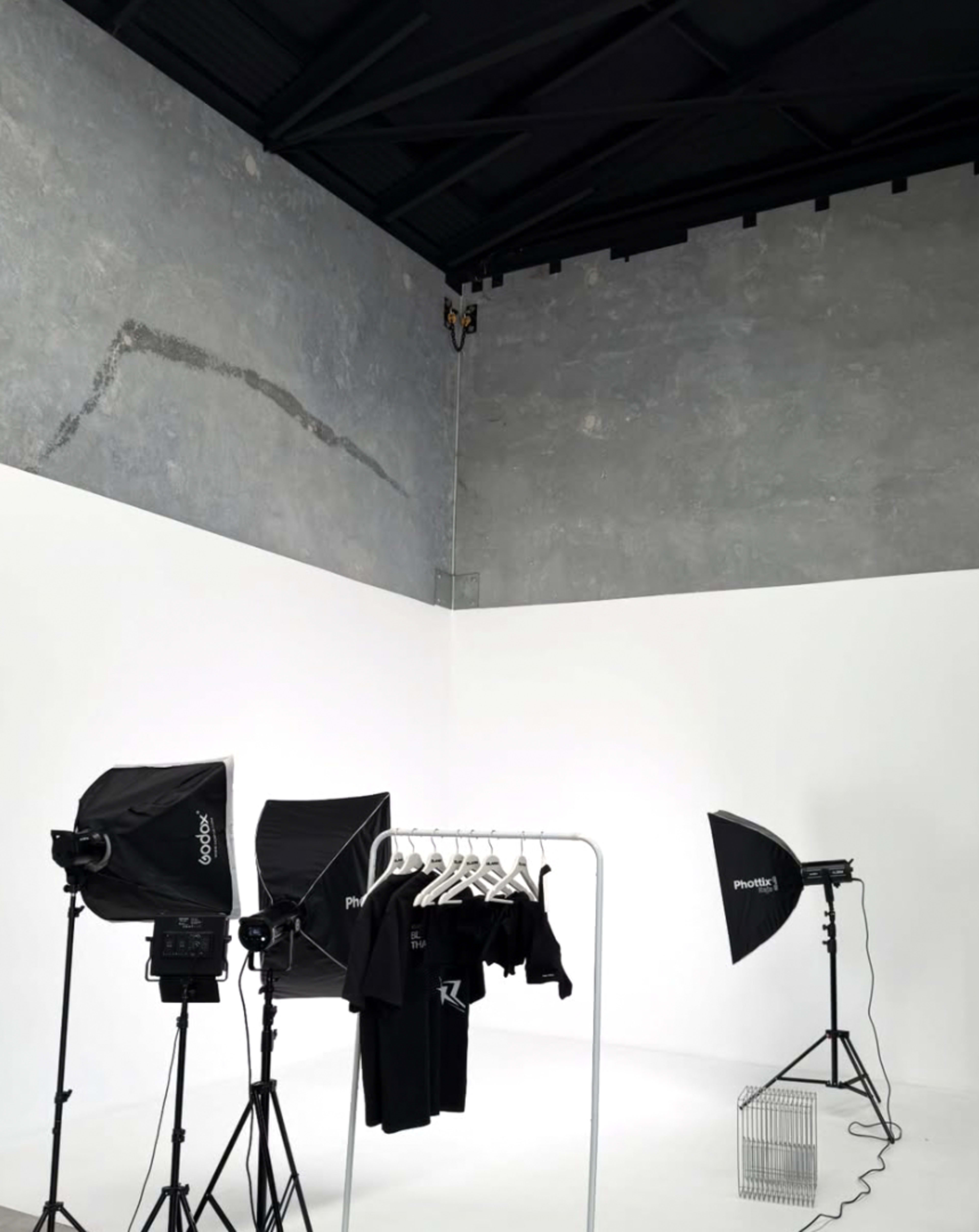 The image shows a minimalist photo studio featuring a clothing rack with black t-shirts, two light setups, and a wire basket against a gray wall.