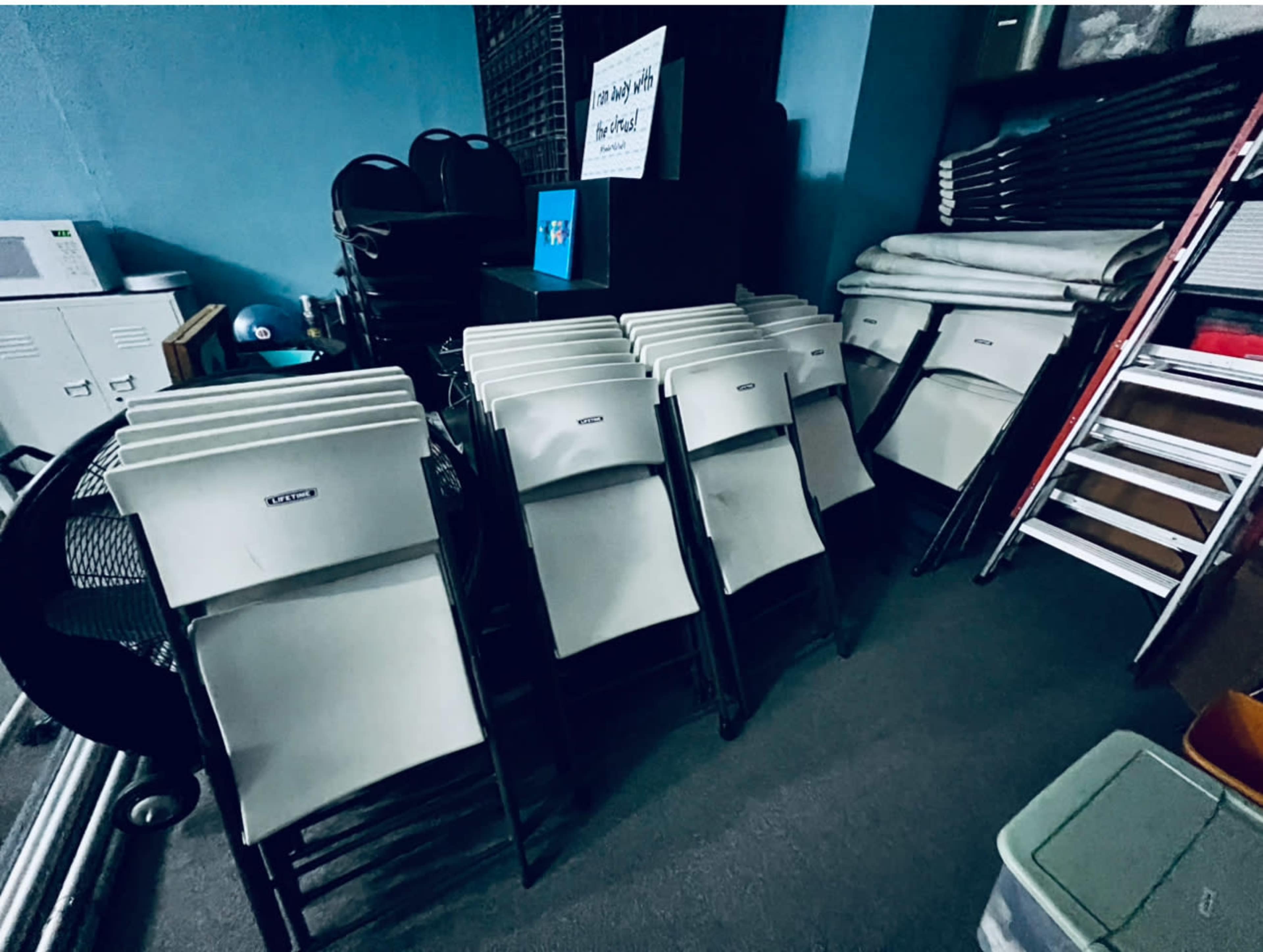 The image shows a stack of several folding chairs arranged against a wall in a room with various storage items, including a ladder and boxes.