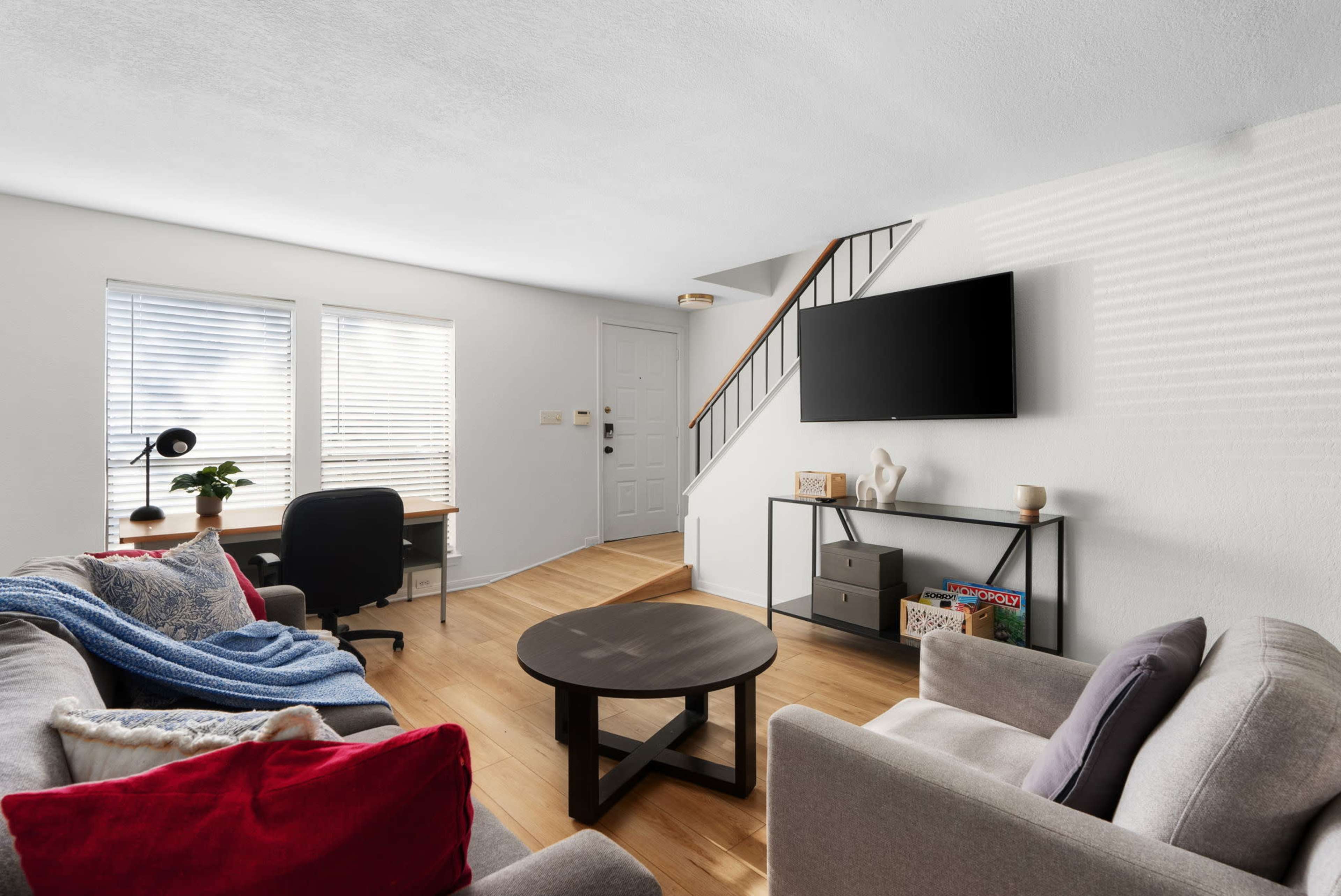 The image depicts a living room with two sofas, a coffee table, a desk, and a television mounted on the wall, alongside a staircase leading to an upper level.