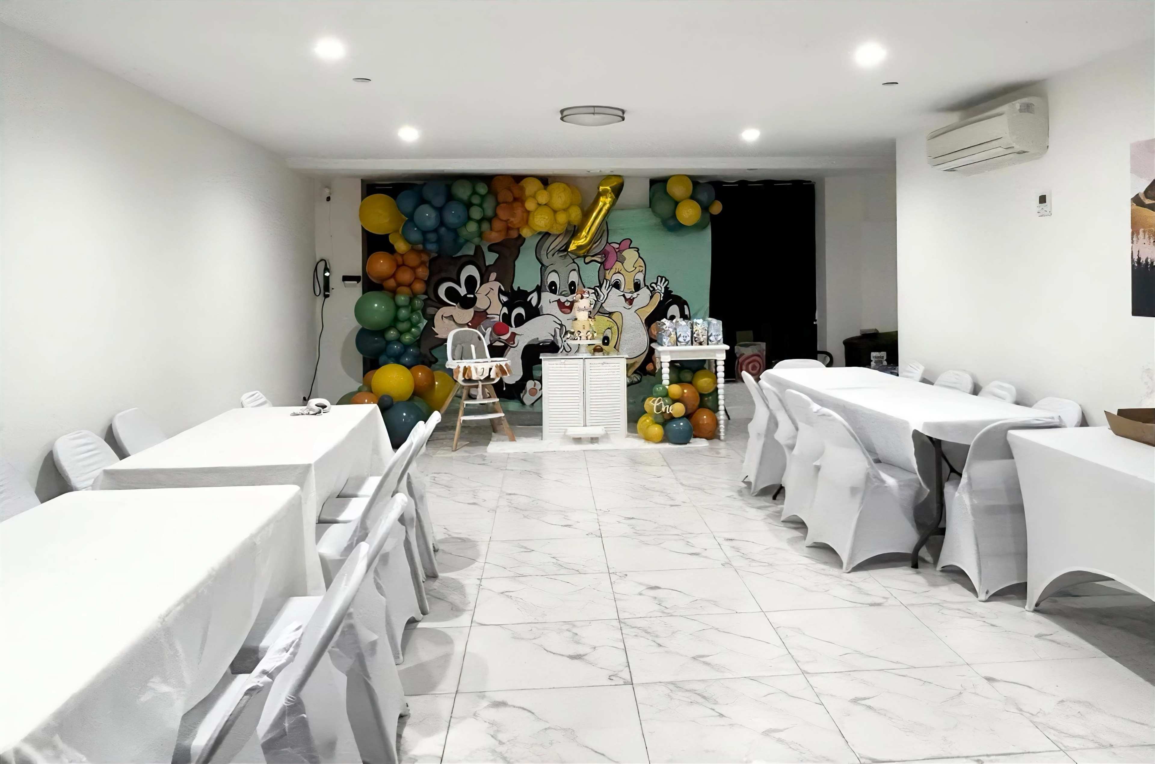 A decorated room with white tables and chairs set up, featuring a colorful balloon wall and a backdrop with cartoon characters.