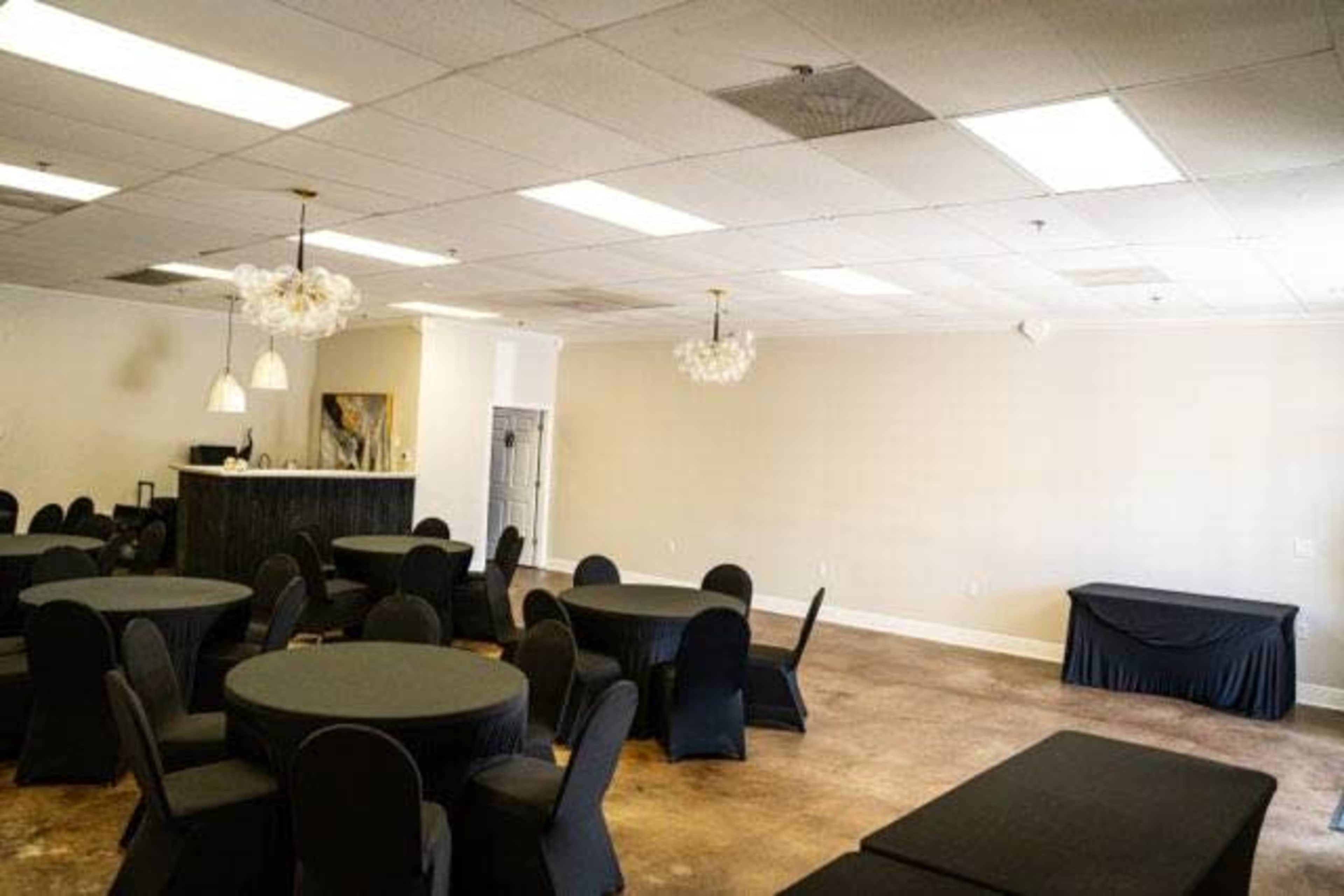 The image shows a spacious event room with several round tables covered in black tablecloths and black chairs, along with a bar area and minimalist decor.