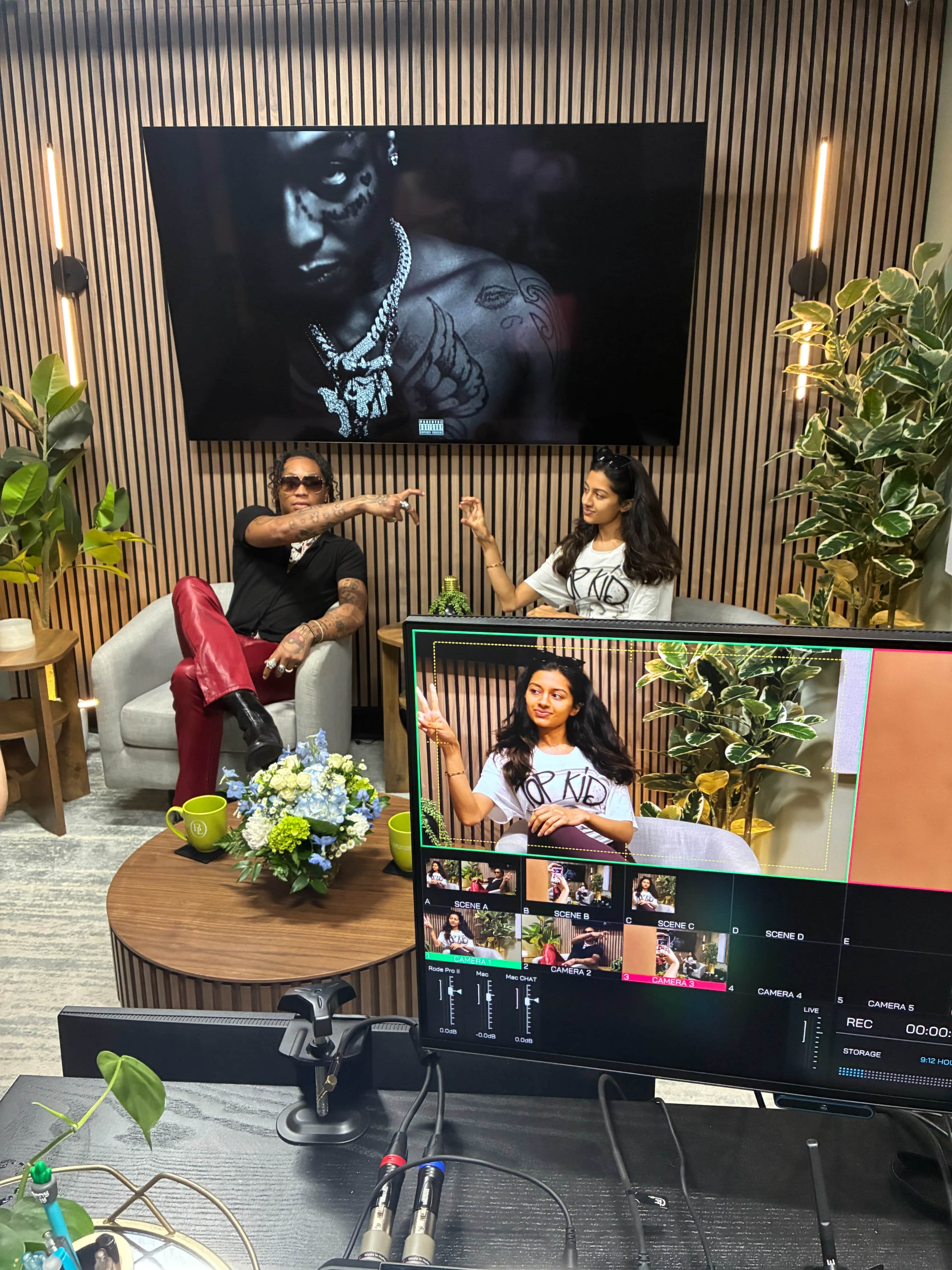 Two individuals sit casually in a studio setting, engaged in a conversation while a camera captures the scene and a large screen displays a visual behind them.