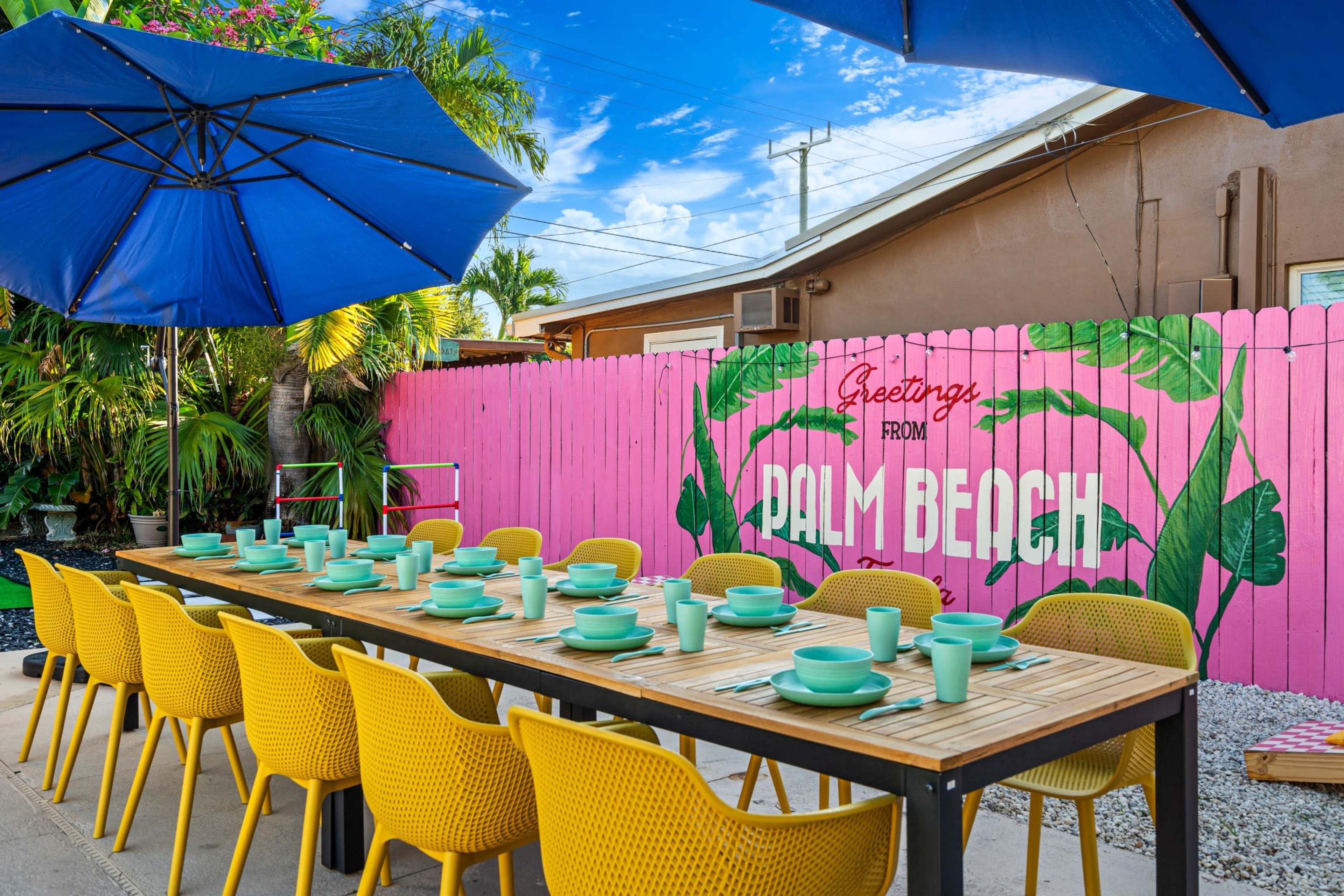 A long dining table with teal plates and white cups is set up under a blue umbrella in front of a pink fence that features a mural saying "Greetings from Palm Beach."