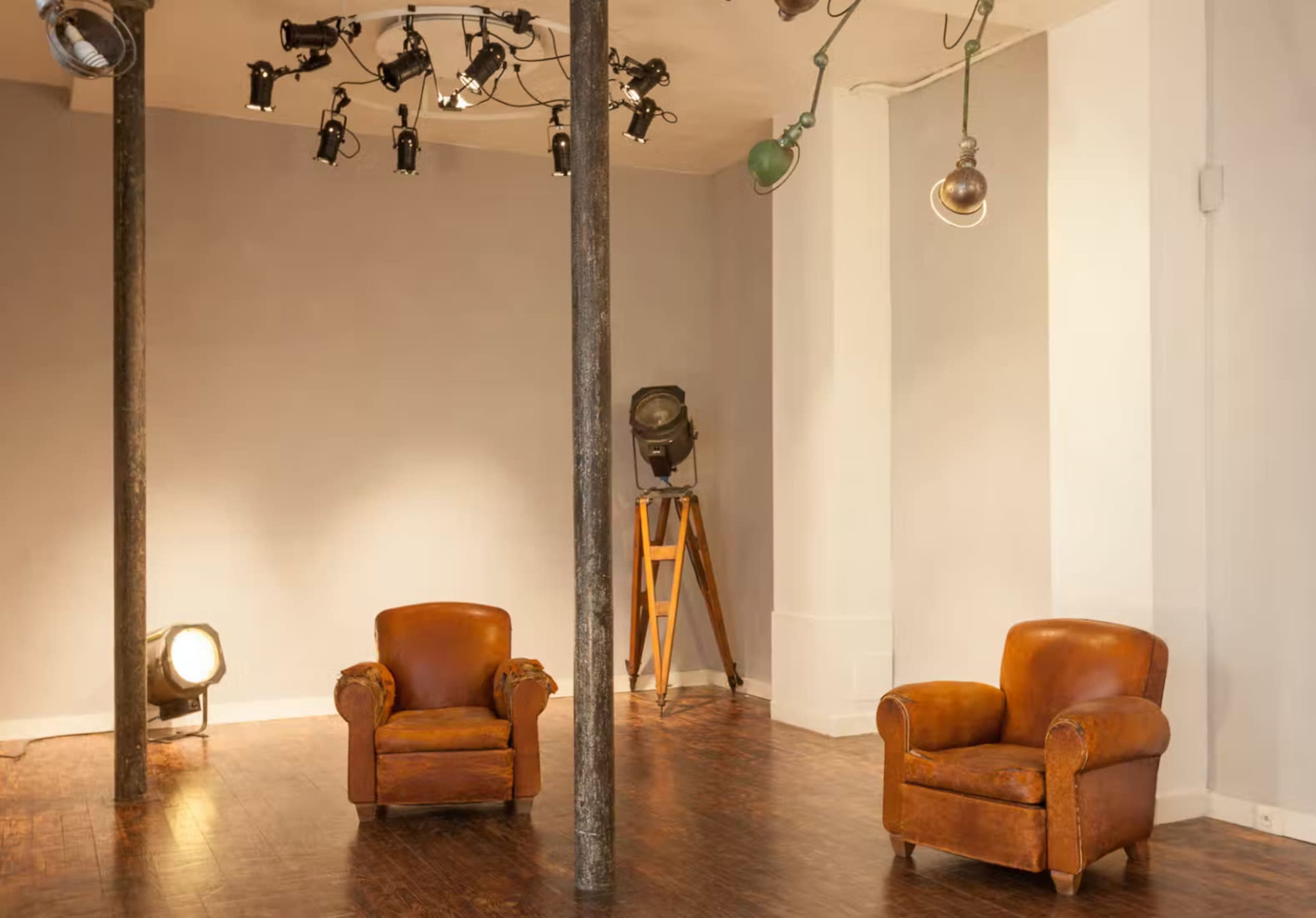 The image shows a minimalist interior space featuring two brown leather armchairs, a tall wooden tripod light stand, and various mounted lights on the ceiling.