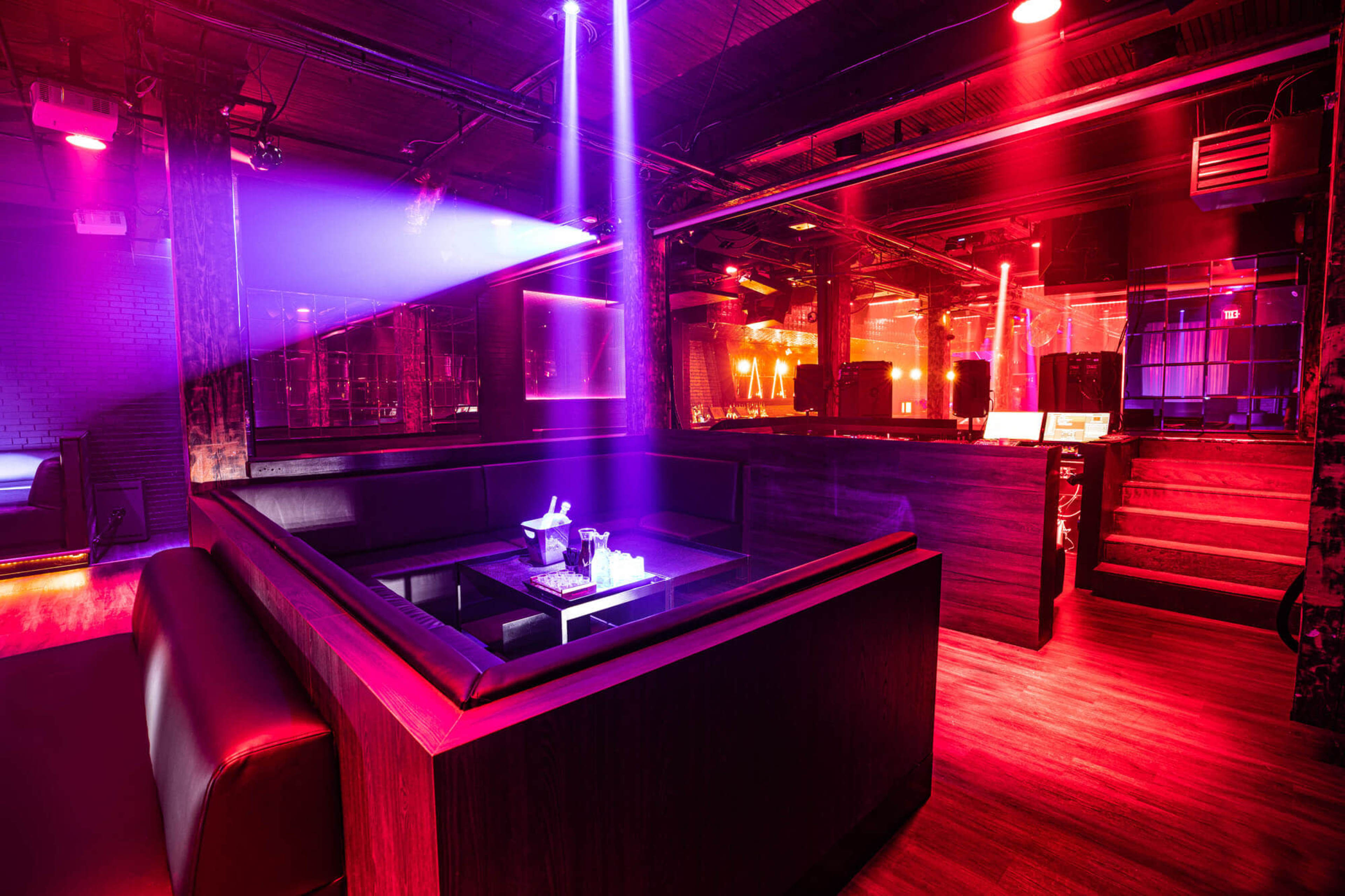 The image shows a dimly lit nightclub interior featuring a seating area with a table, illuminated by colorful neon lights.