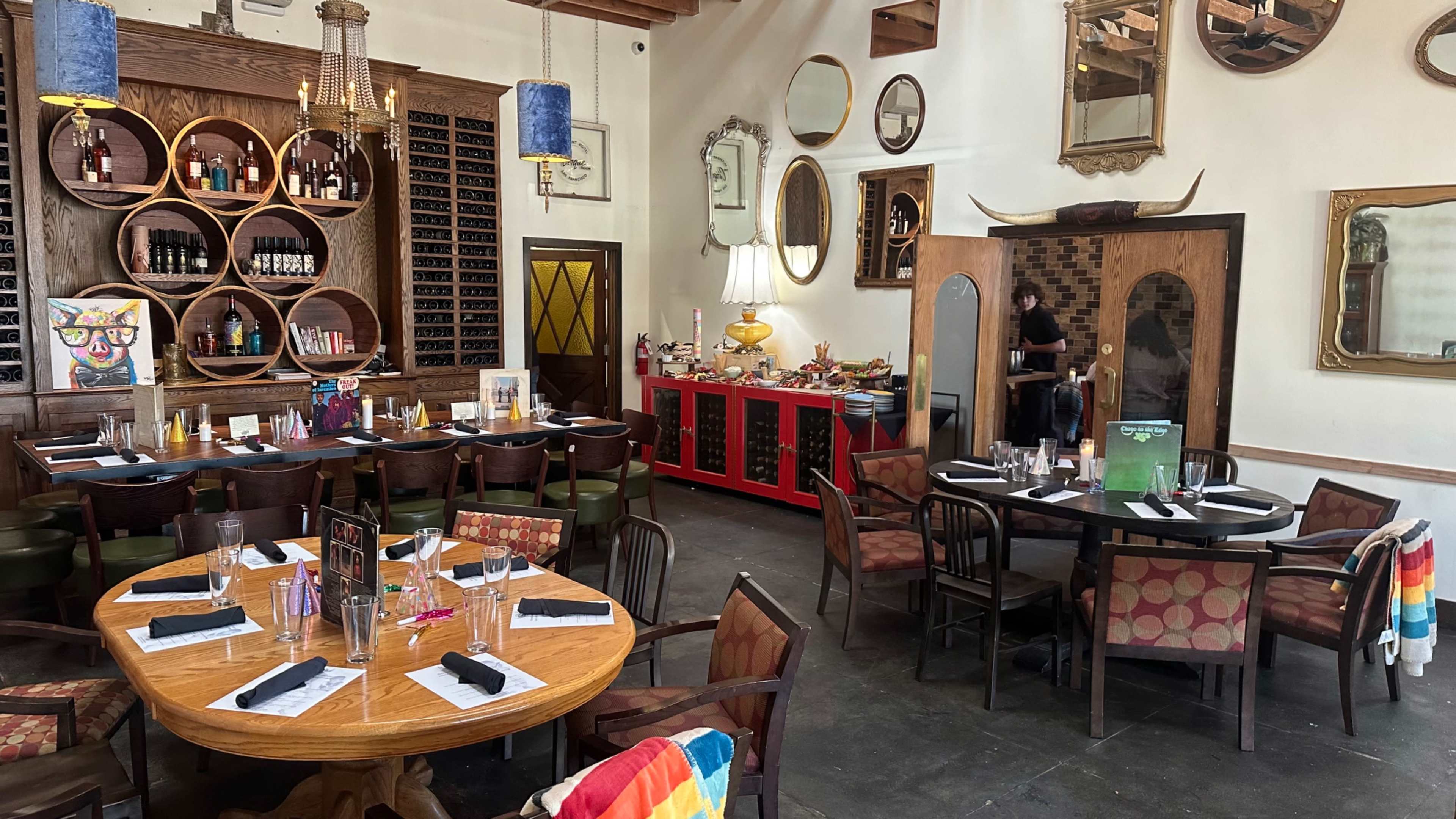 The image shows a restaurant interior with wooden tables set for dining, a wine rack on the wall, circular mirrors, and a colorful buffet counter.