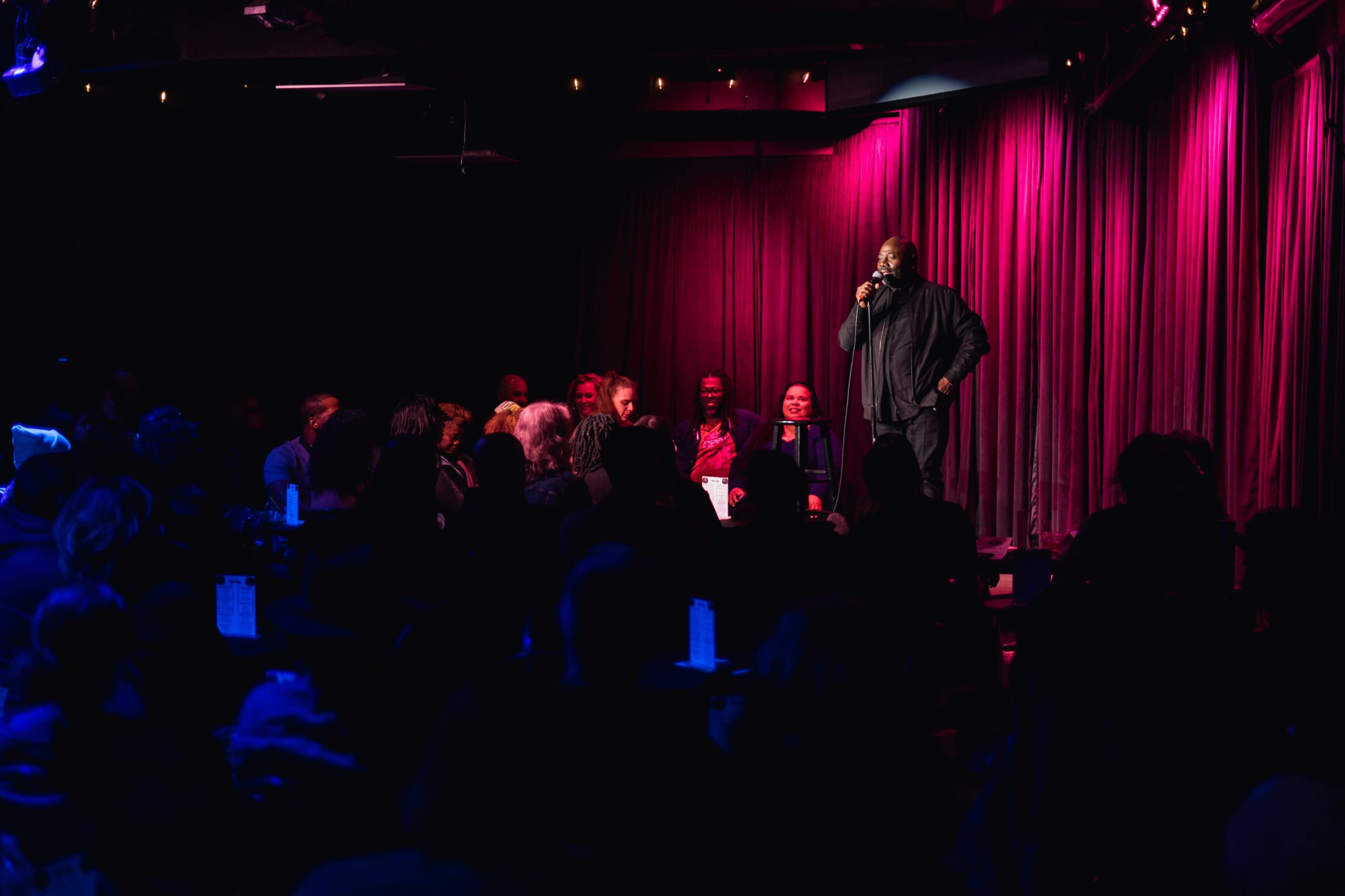 A comedian stands on stage in front of an audience at a dimly lit comedy club with pink and purple lighting.