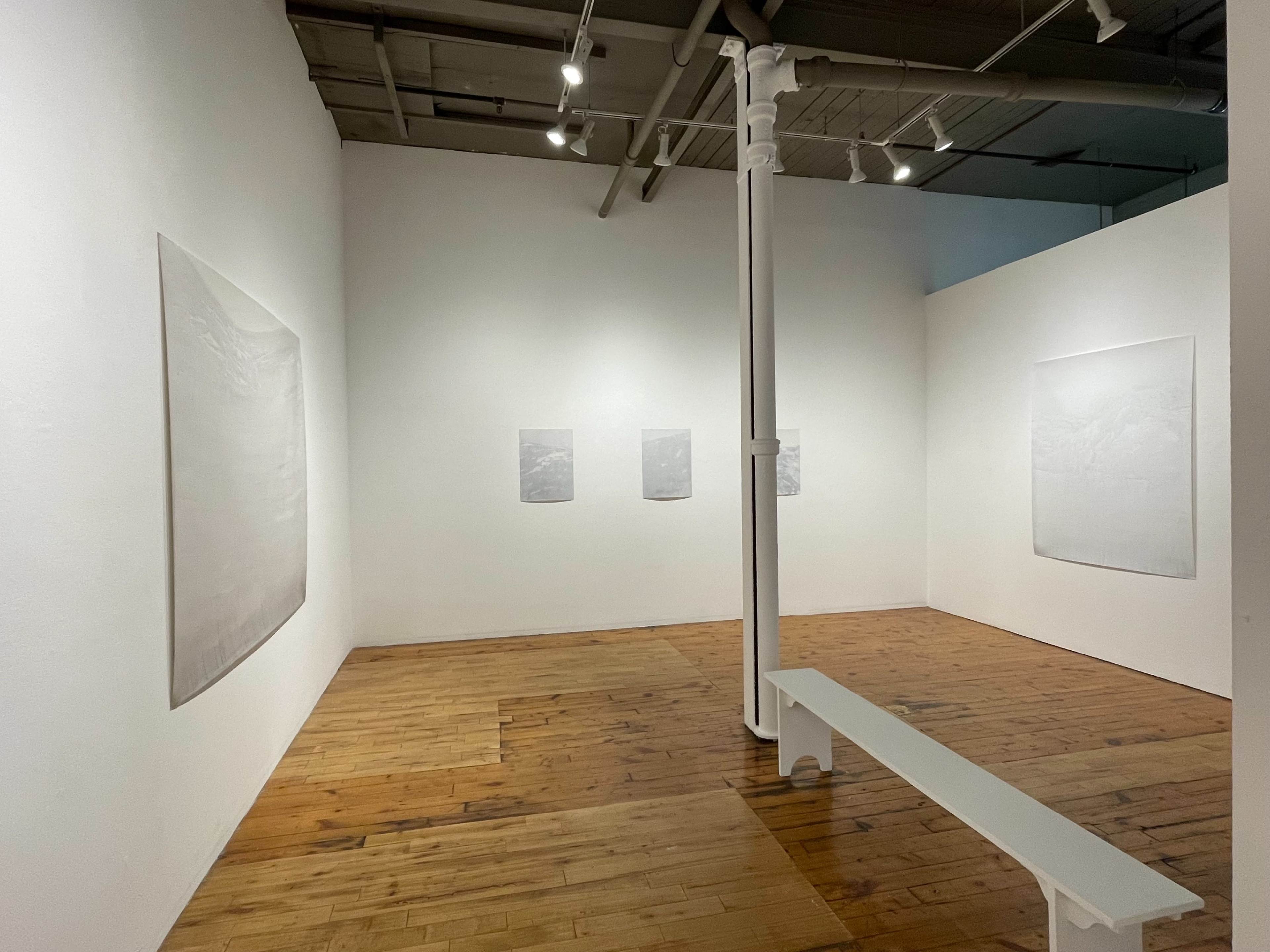 An art gallery with white walls, wooden flooring, and a white bench in the center, featuring several minimalist artworks displayed on the walls.
