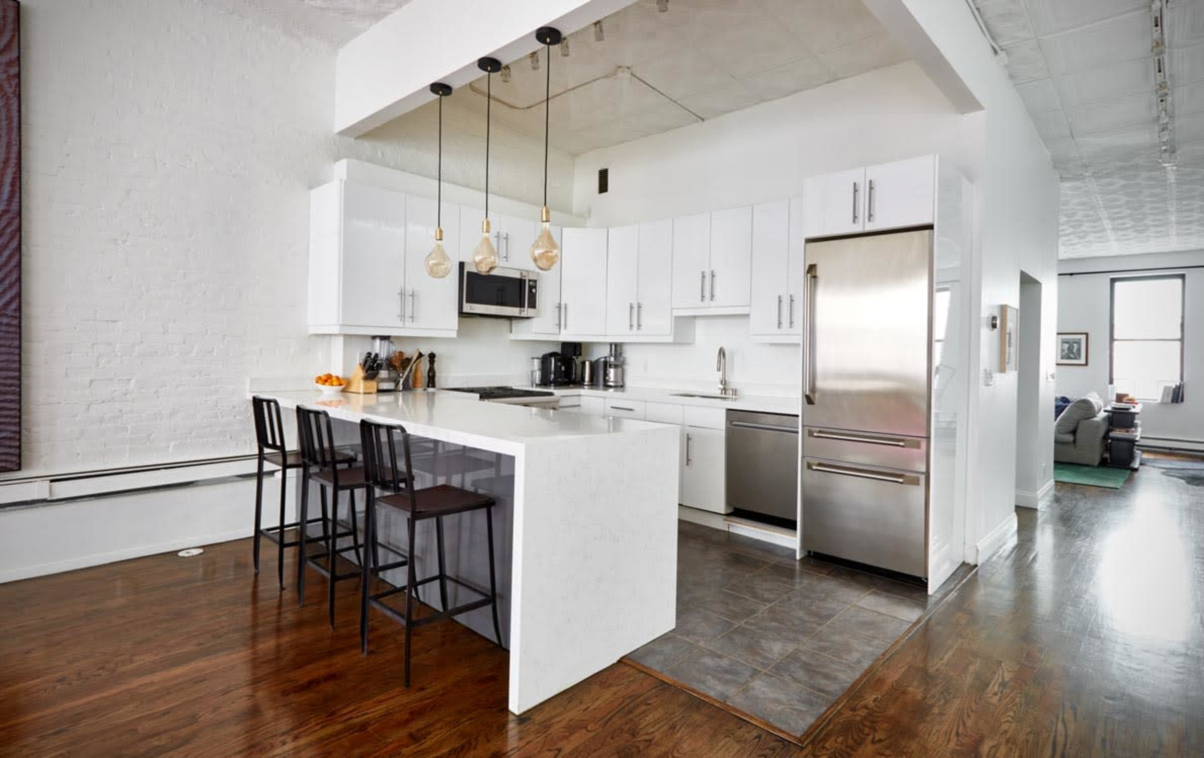 The image shows a modern kitchen featuring white cabinetry, stainless steel appliances, and a small island with three black stools.