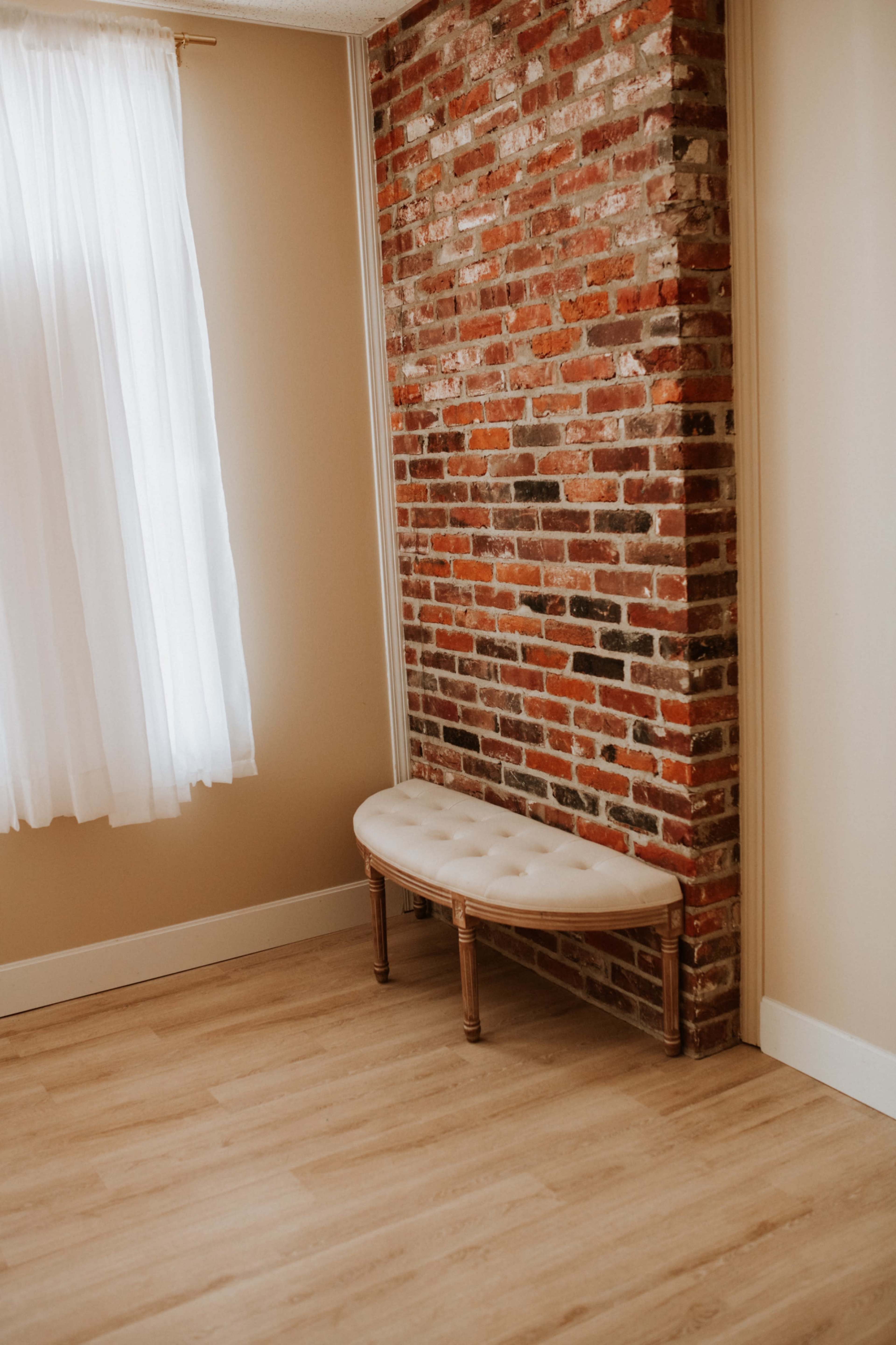 The image shows a small, light-colored room with a brick accent wall and a cushioned bench against it.