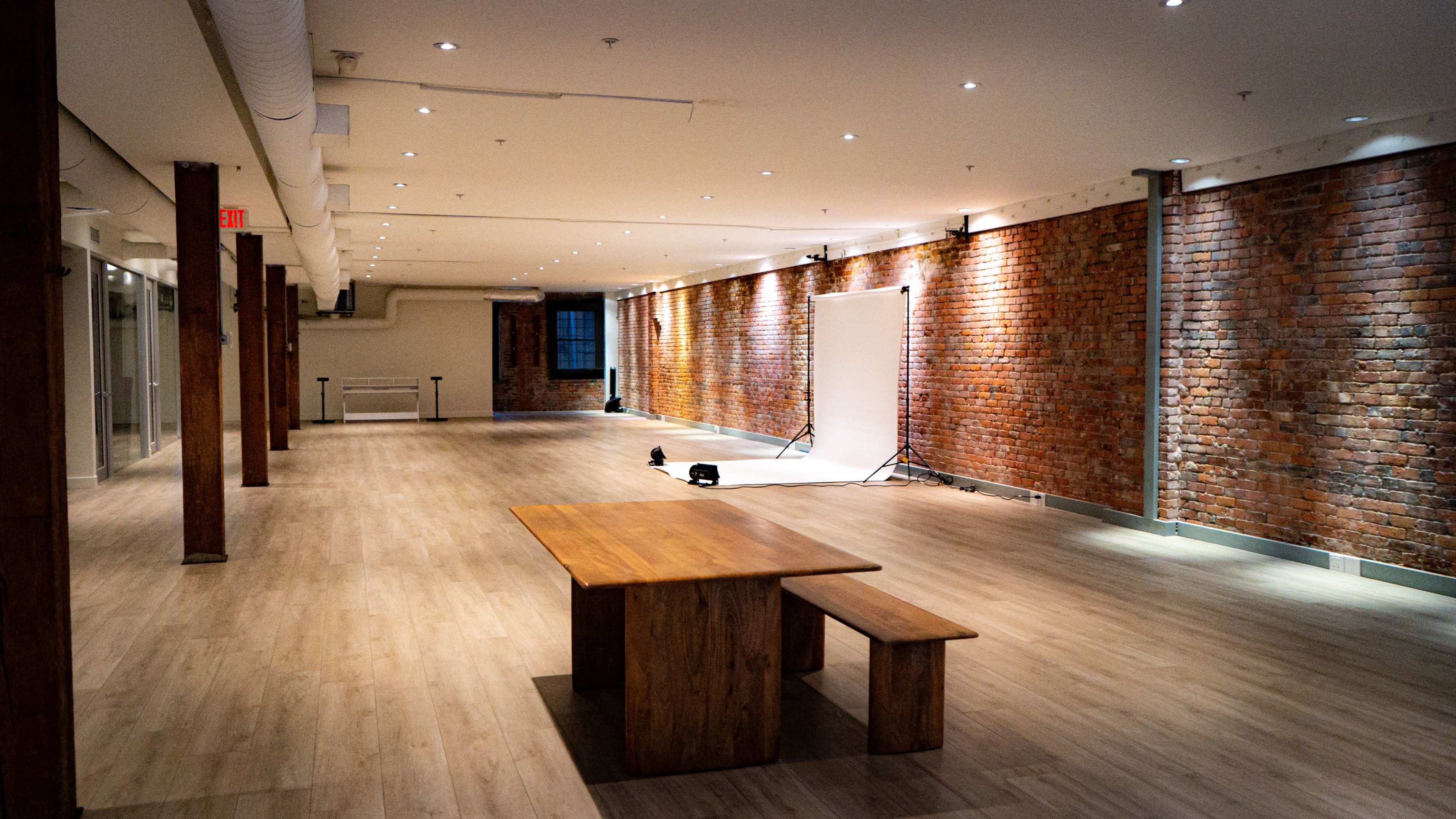 The image shows a spacious interior of a venue with wooden flooring, exposed brick walls, and a simple wooden table and bench set in the center.