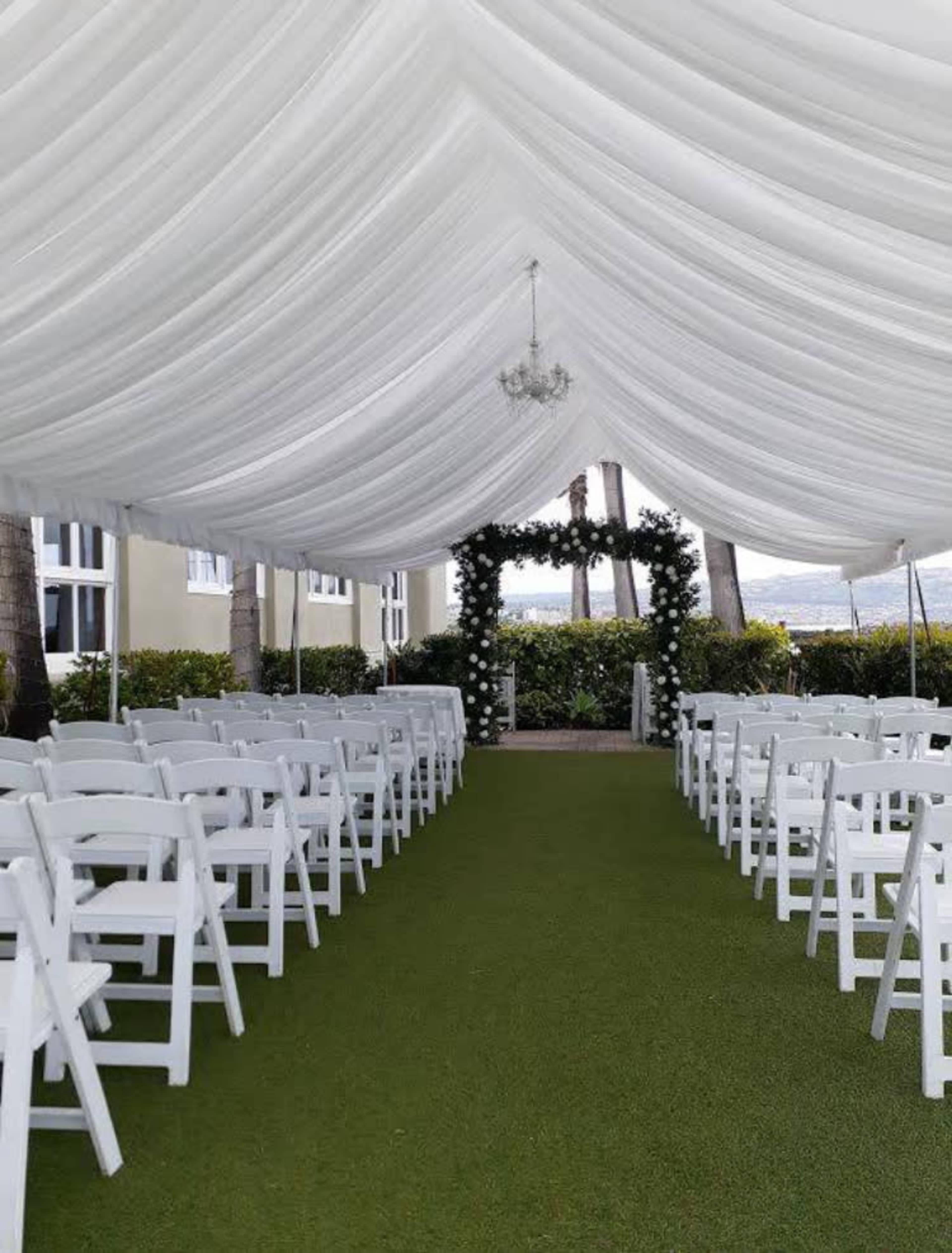 A tented outdoor venue features rows of white chairs set on green grass, leading to an arch adorned with greenery.