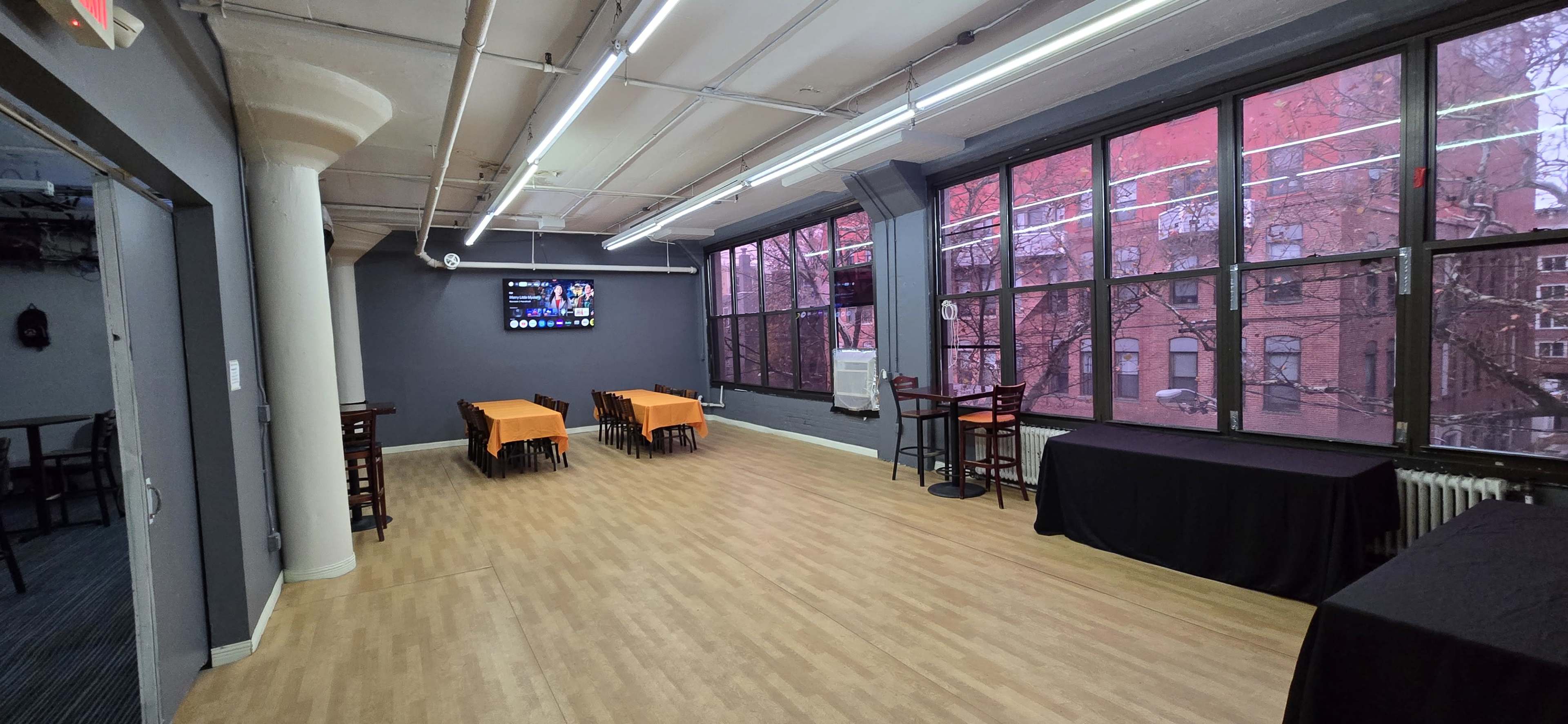 The room features a spacious layout with large windows, two tables with orange covers, and a black table along one wall.