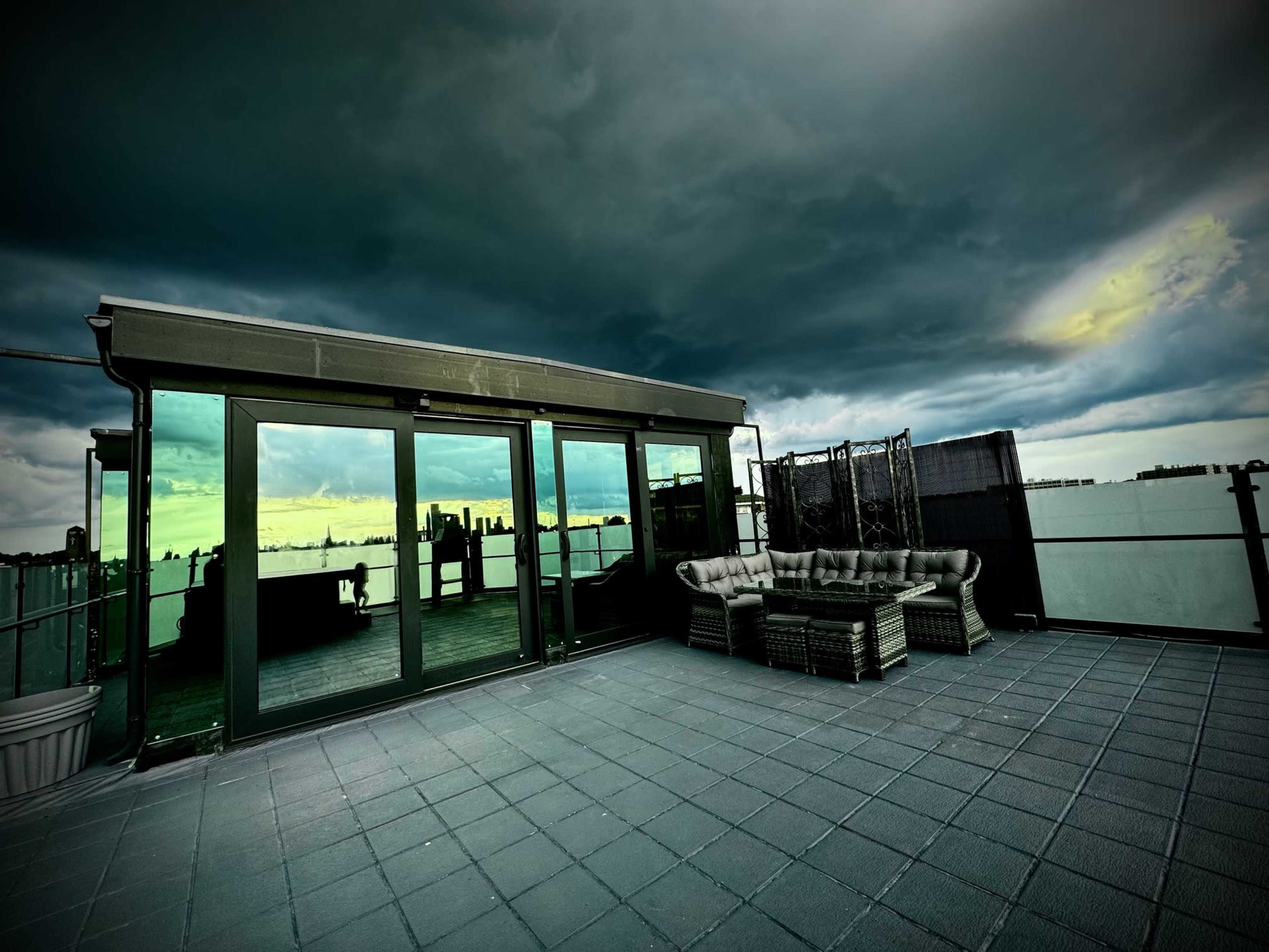 A spacious rooftop terrace features large glass doors and modern outdoor furniture, set against a dramatic, overcast sky.