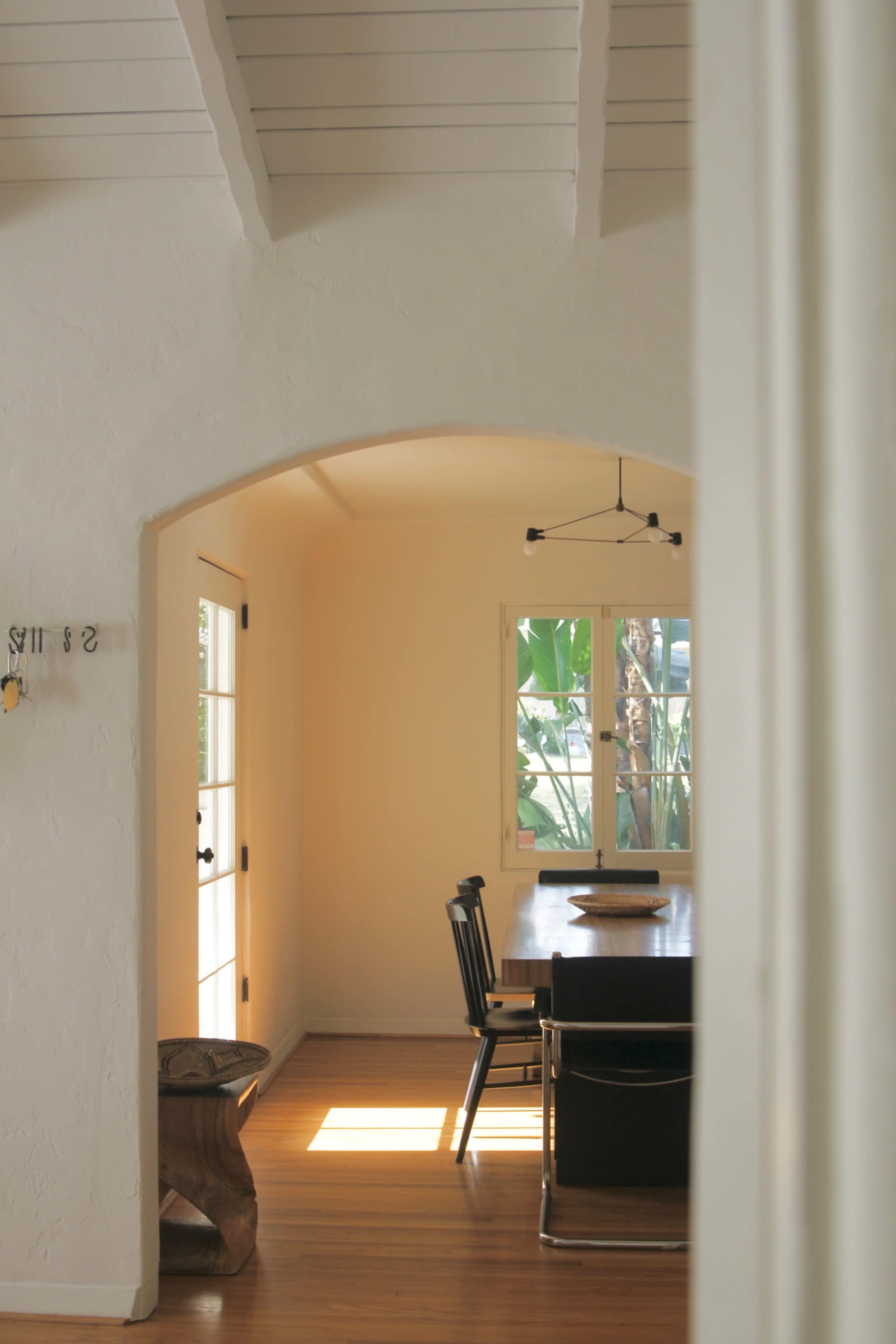 A light-filled dining area features a wooden table and black chairs beneath a modern light fixture, framed by an arched doorway and visible plants outside.