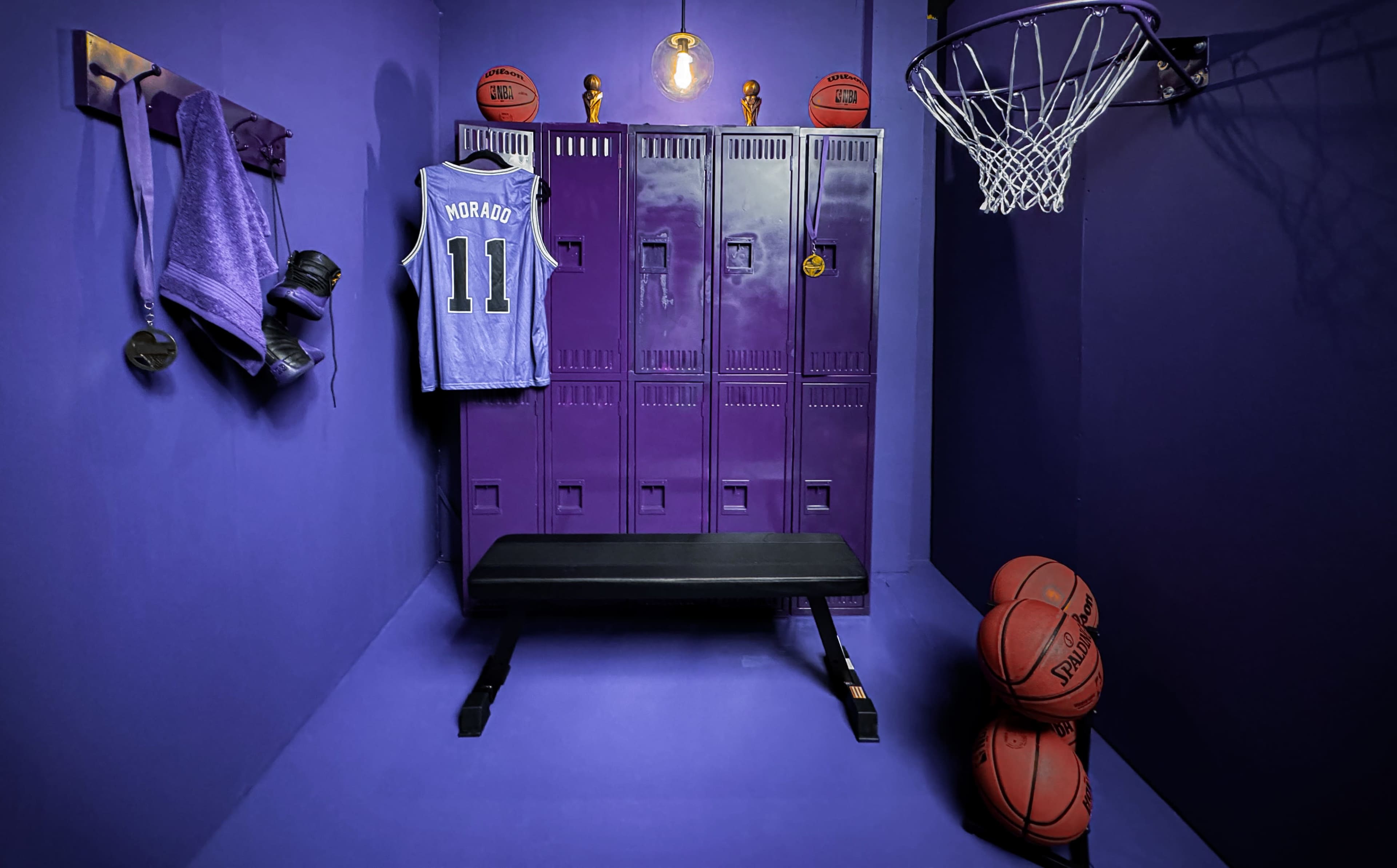 A purple-themed locker room with a bench, basketball jerseys hanging on the wall, and stacked basketballs near a hoop.