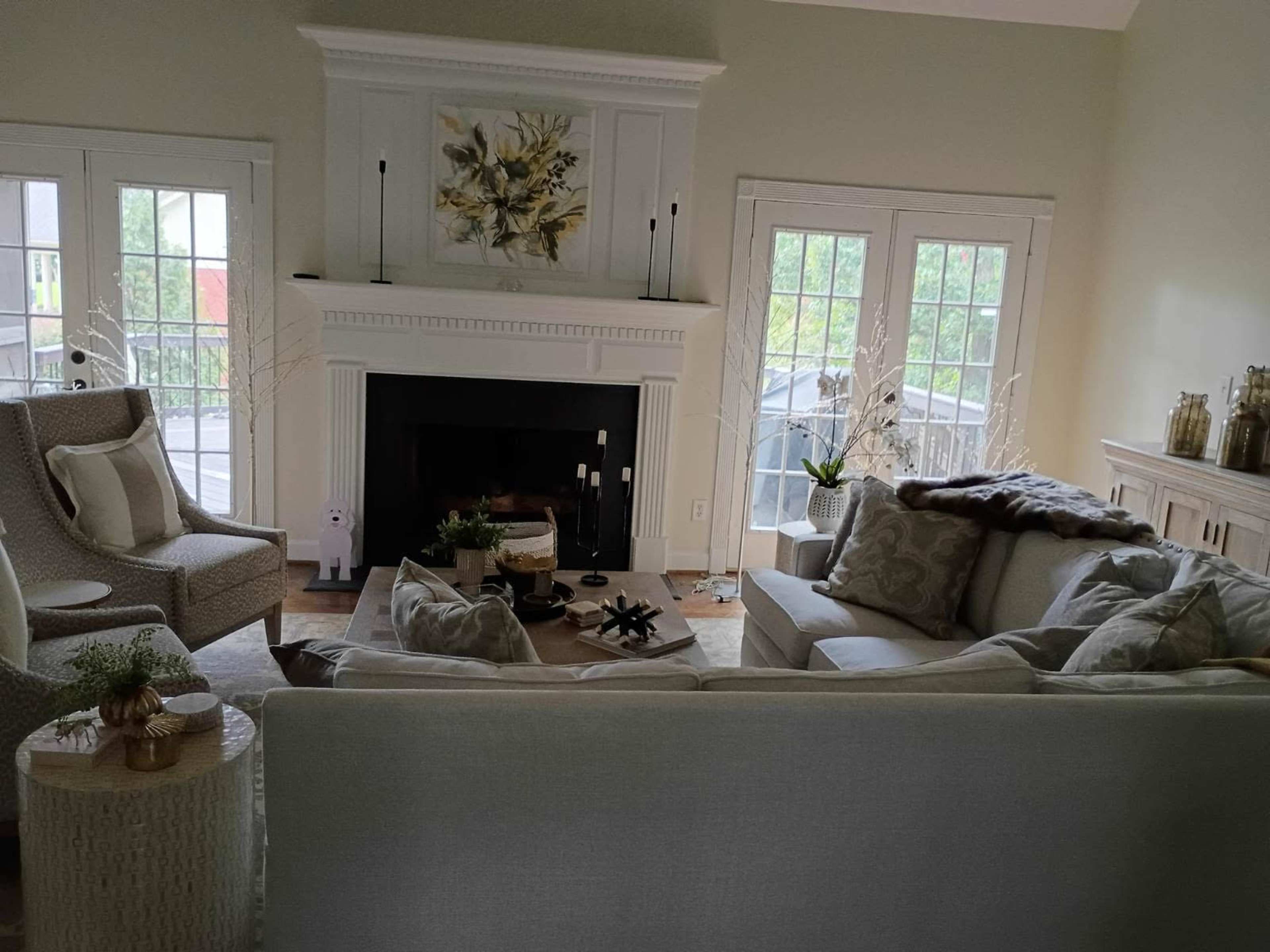 The image shows a cozy living room with light-colored furniture, a fireplace, and large windows letting in natural light.