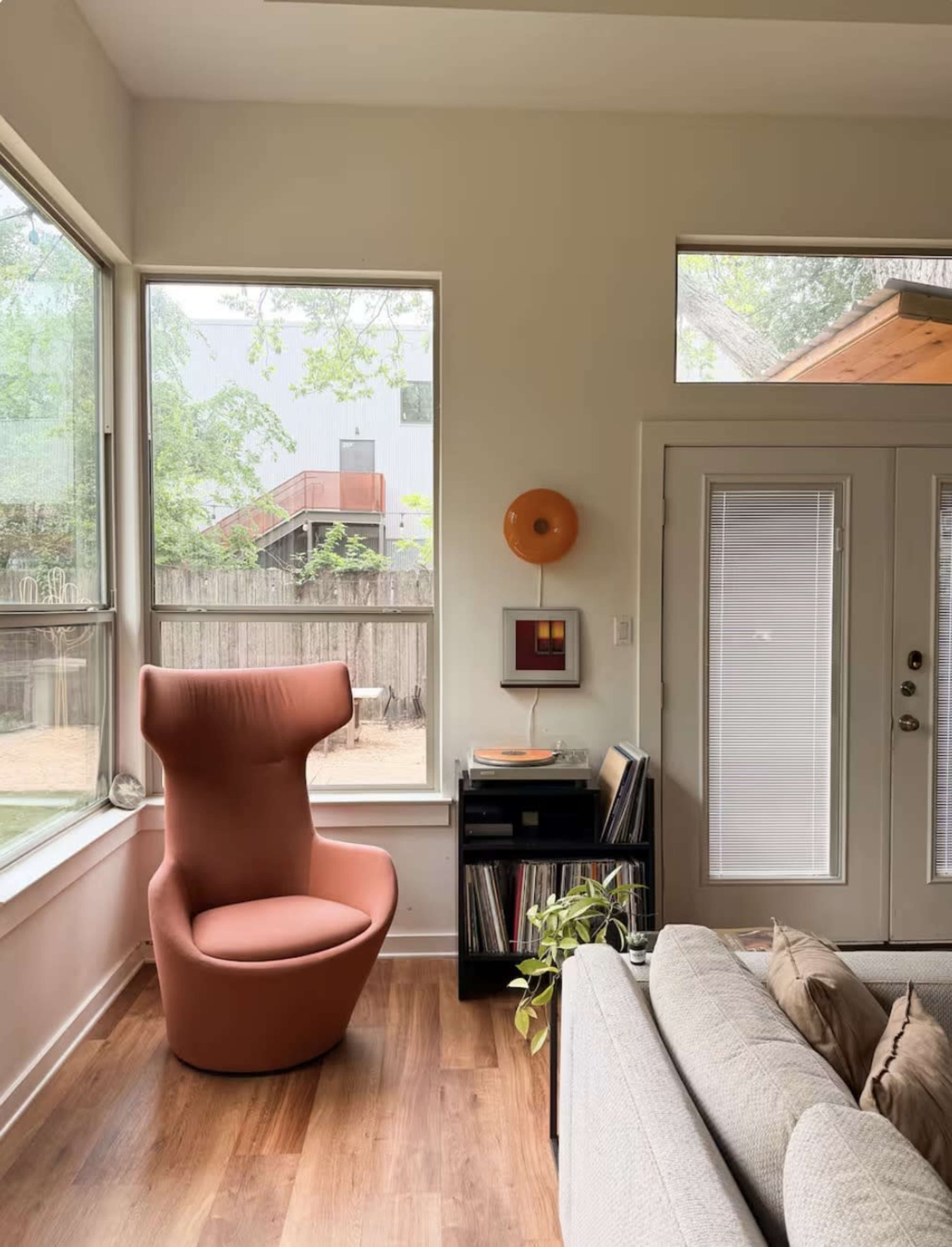 The image shows a cozy living room area featuring a unique, curved orange chair next to a small shelving unit with vinyl records, large windows allowing natural light, and a door leading outside.