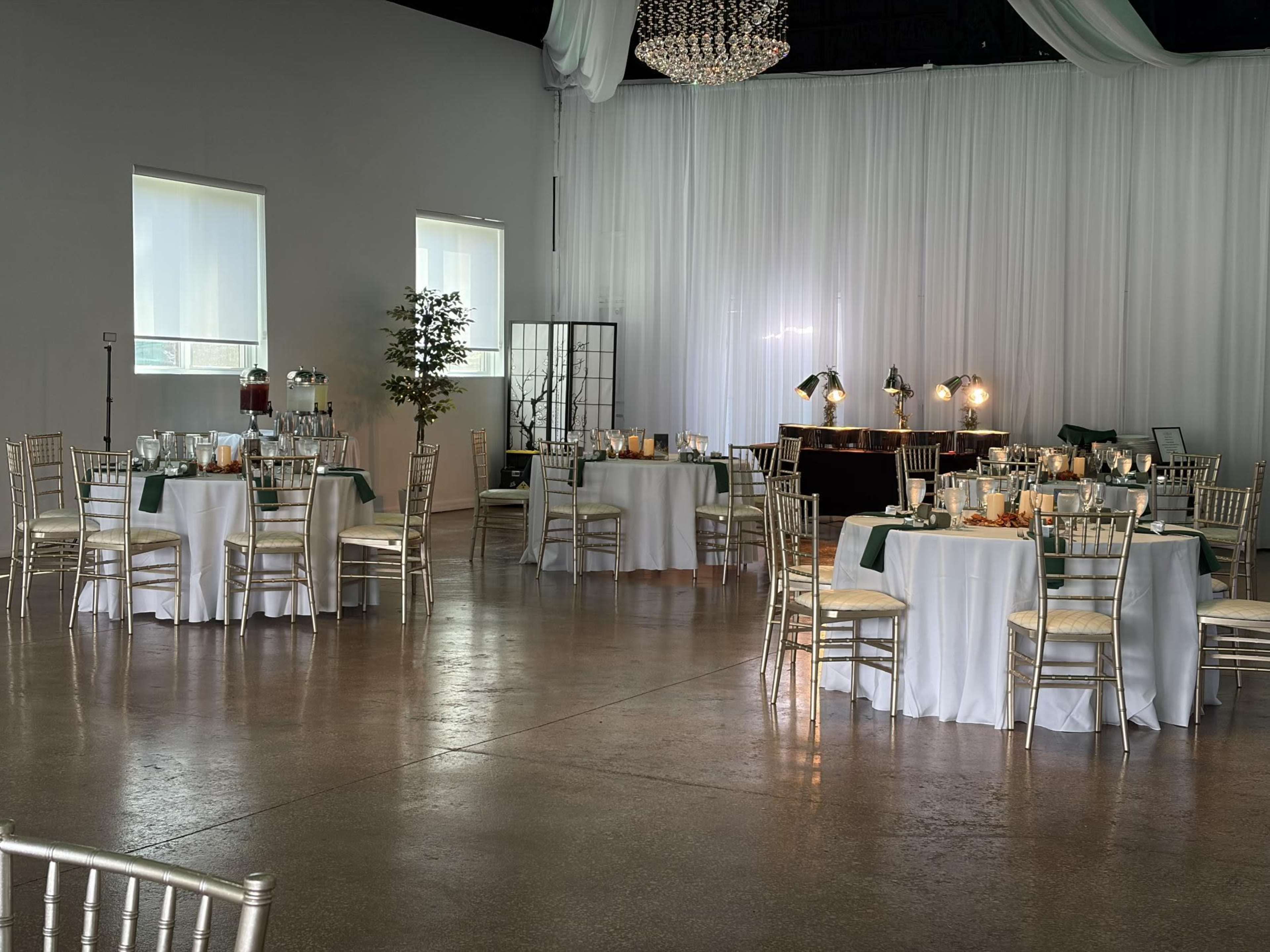 The image shows a banquet hall setup with several round tables elegantly arranged, illuminated by soft lighting and decorated with white tablecloths and green accents.