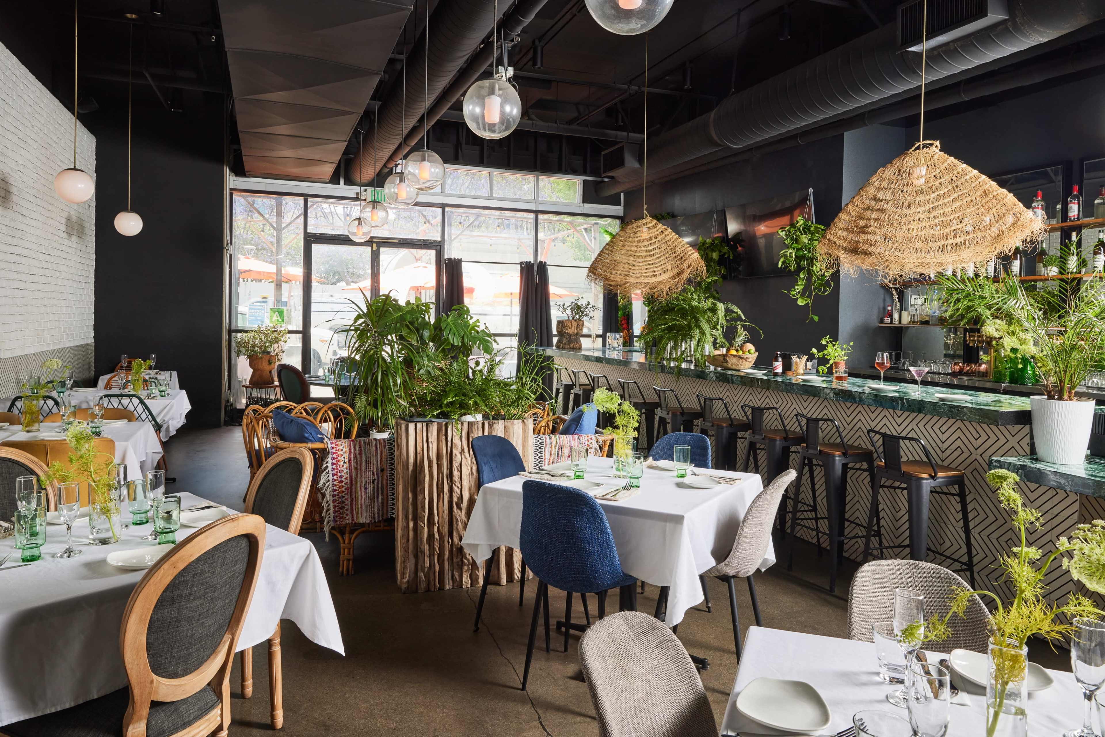 The image shows a modern restaurant interior featuring multiple tables with white tablecloths, a green bar area, and hanging woven light fixtures surrounded by potted plants.