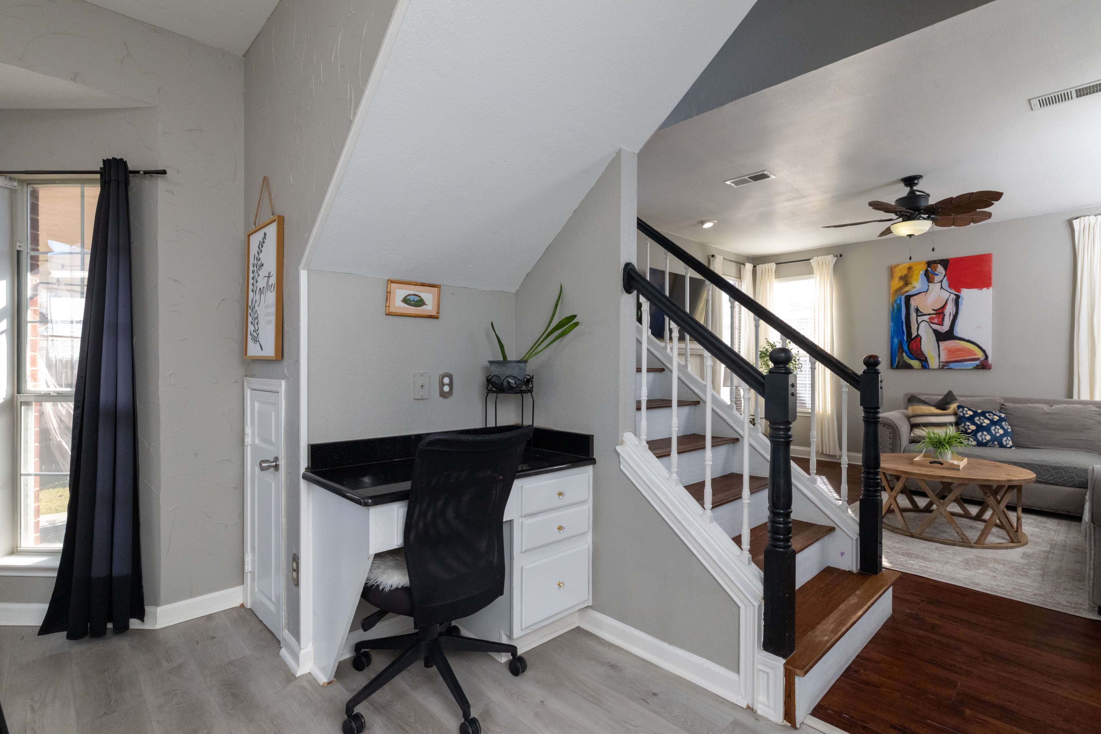 The image shows a small home office nook with a black chair and desk situated near a staircase, adjacent to a living room that features a gray sofa and colorful artwork.