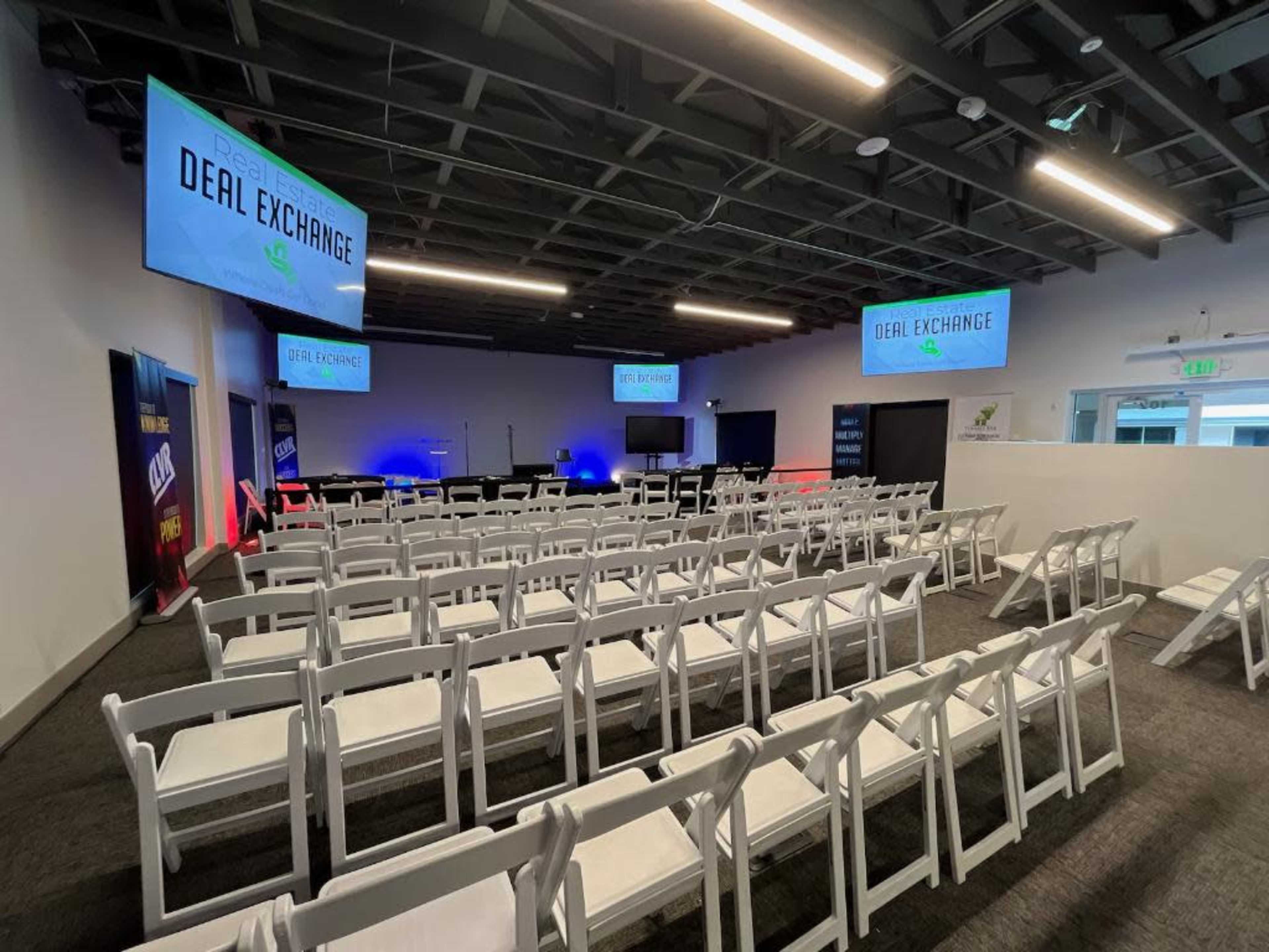 A conference room setup with multiple rows of white chairs facing a stage area lit with blue and red lighting, and two large screens displaying "DEAL EXCHANGE."