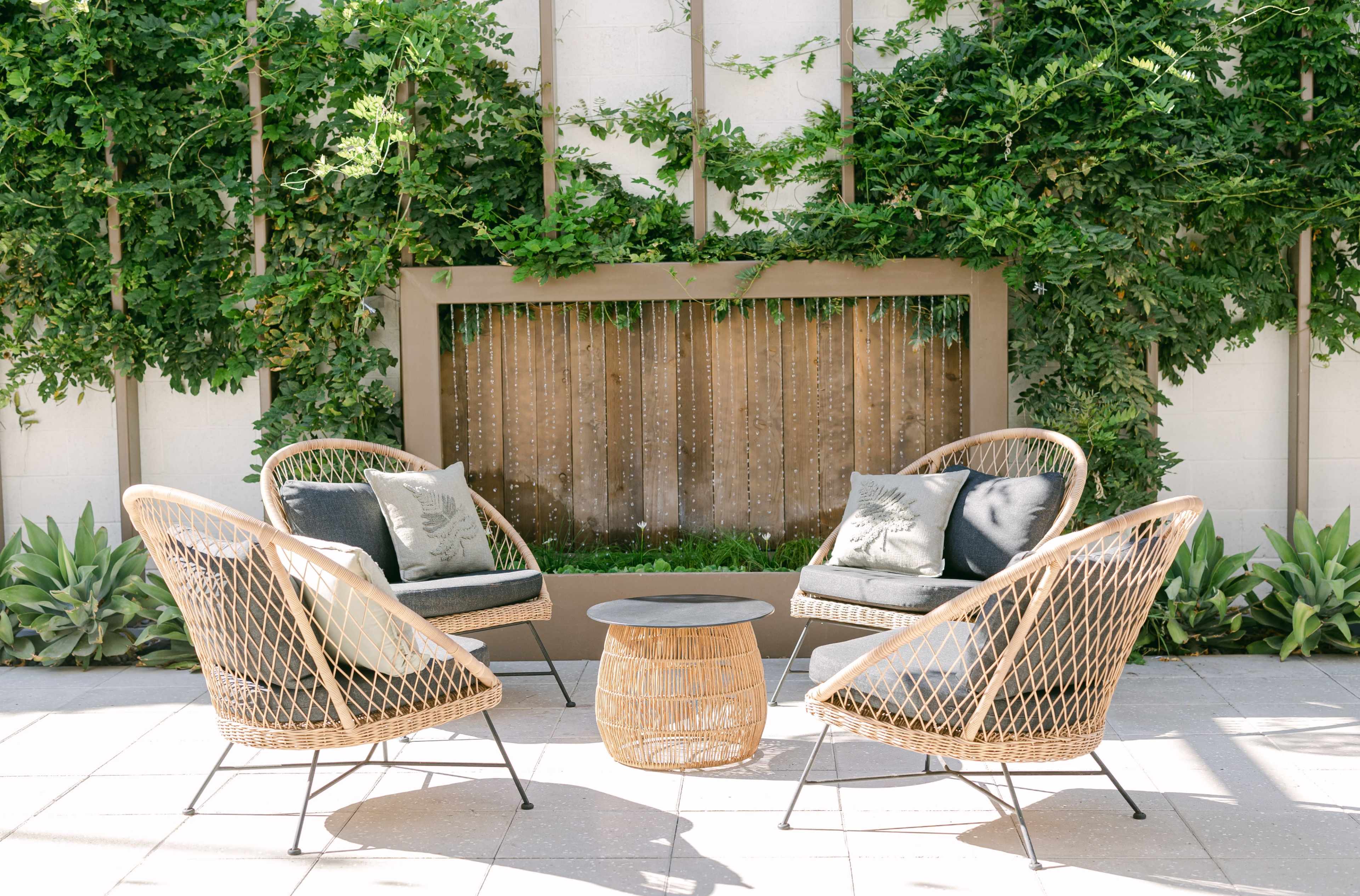 A patio seating area features four woven chairs arranged around a round coffee table, with a backdrop of greenery and a wooden panel.