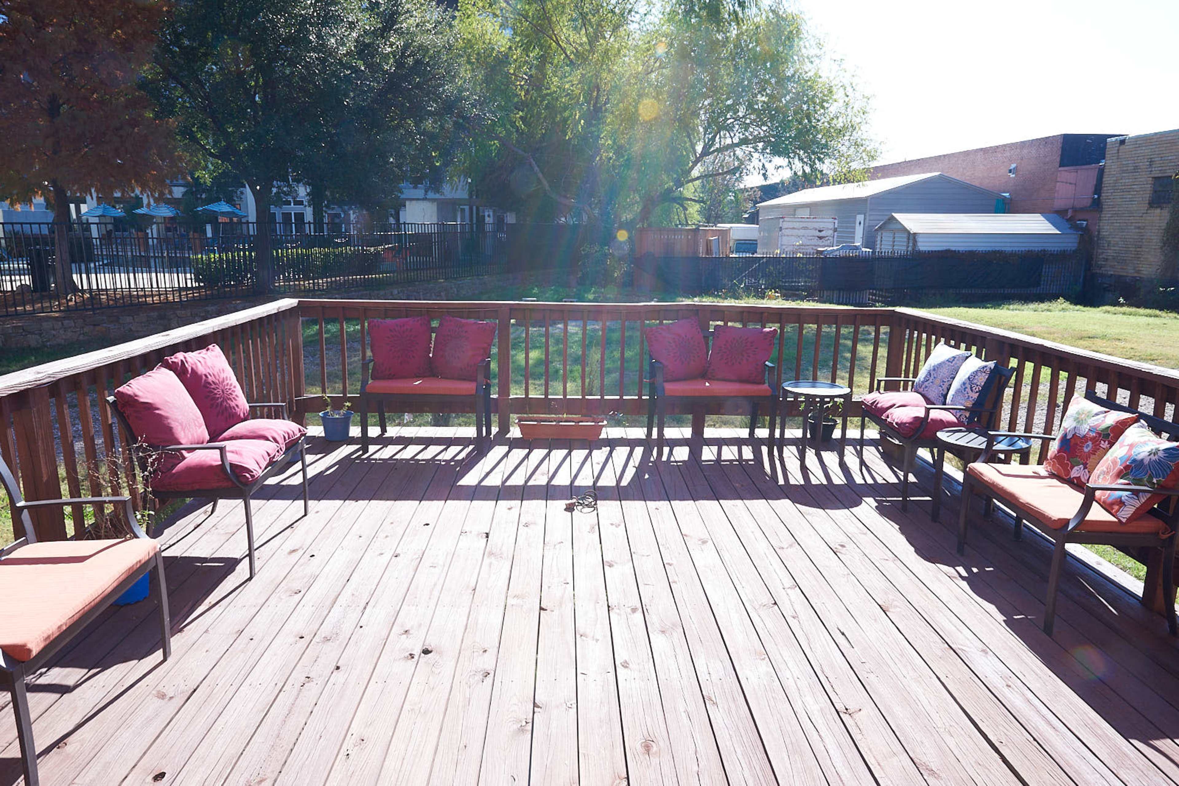 A wooden deck features several chairs and cushions arranged around a central space, with a grassy area visible in the background.