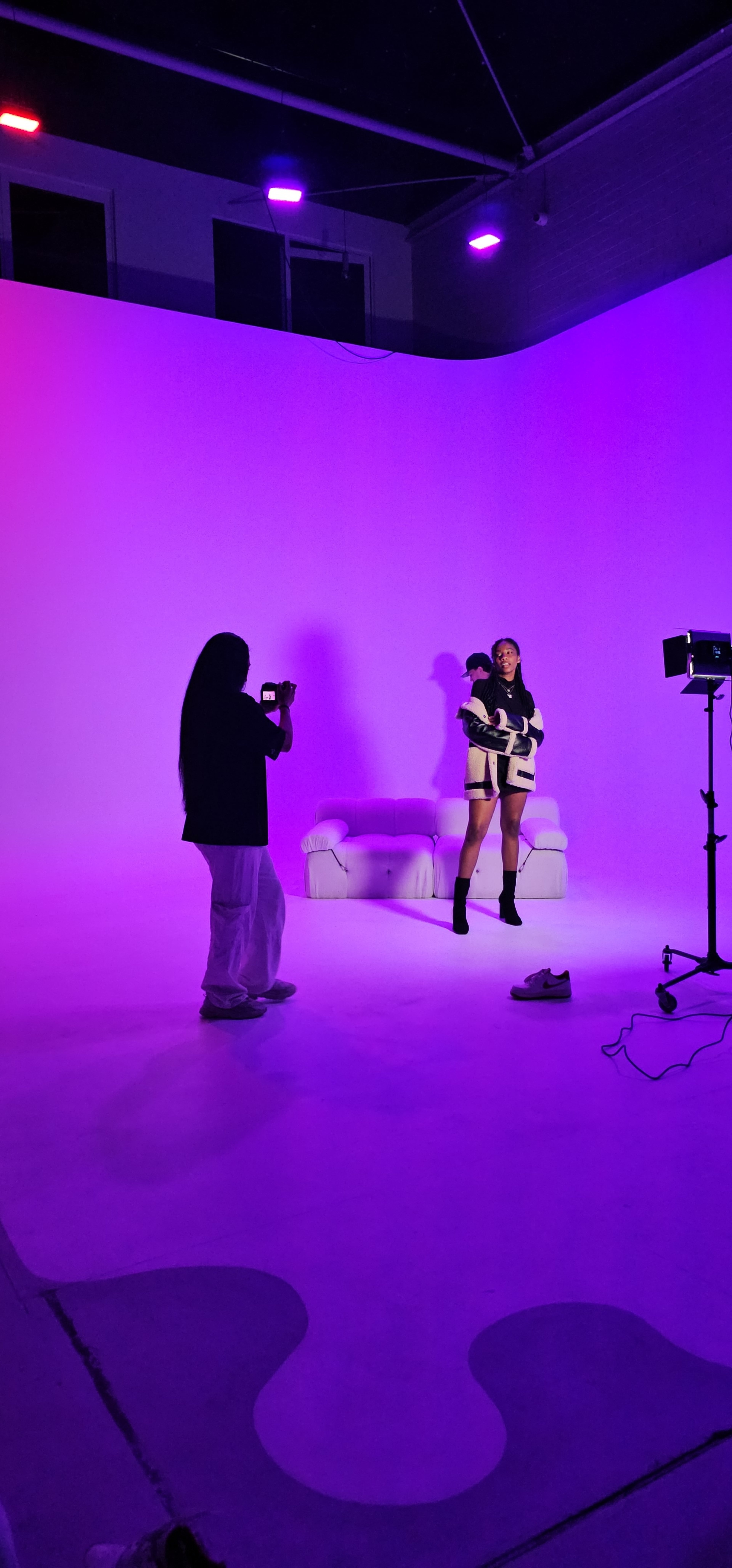 A person is taking photographs of a model posing with a plush armchair against a brightly lit purple backdrop.
