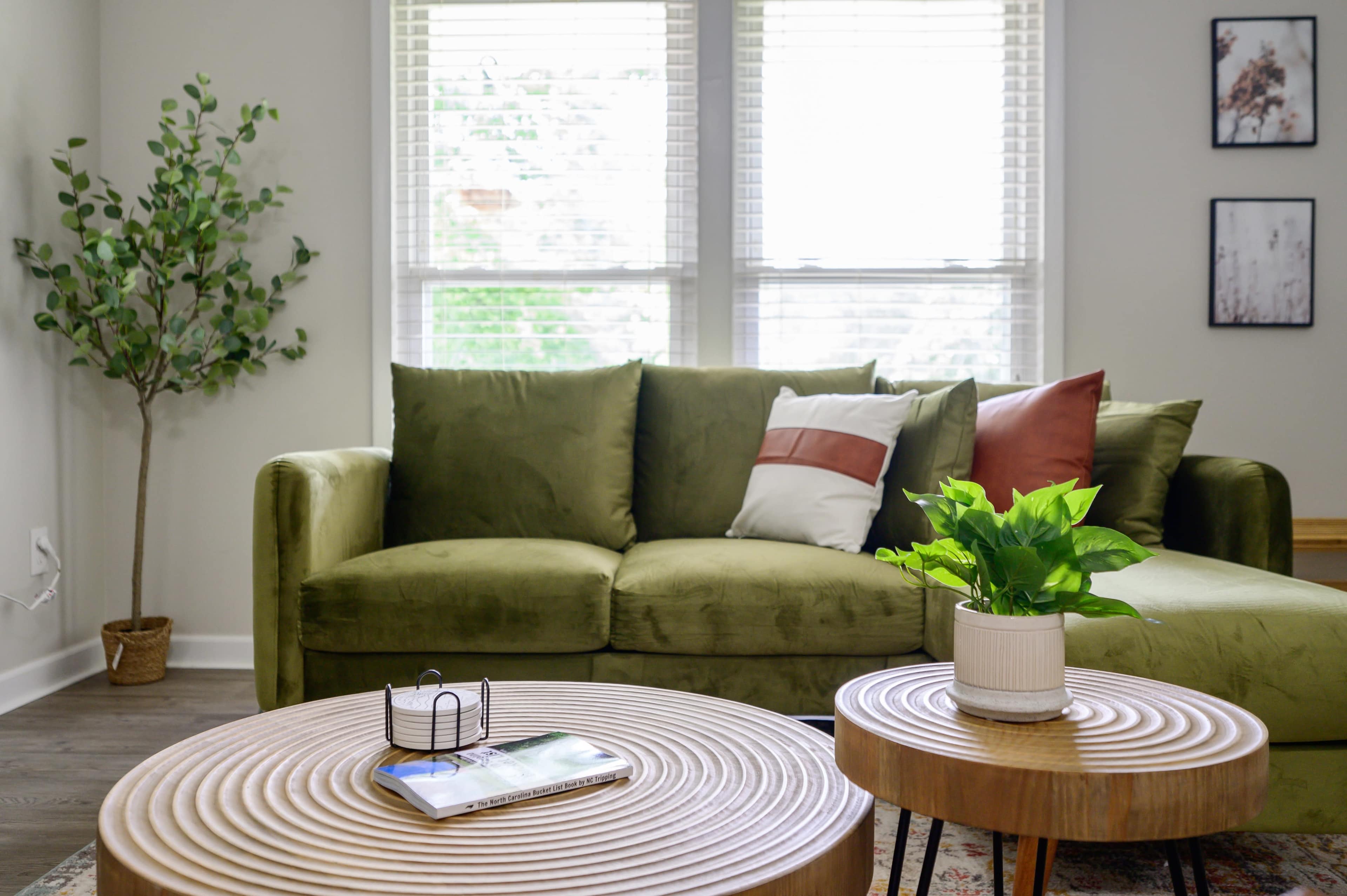 A modern living room with a green couch, two wooden coffee tables, and a potted plant near a window.