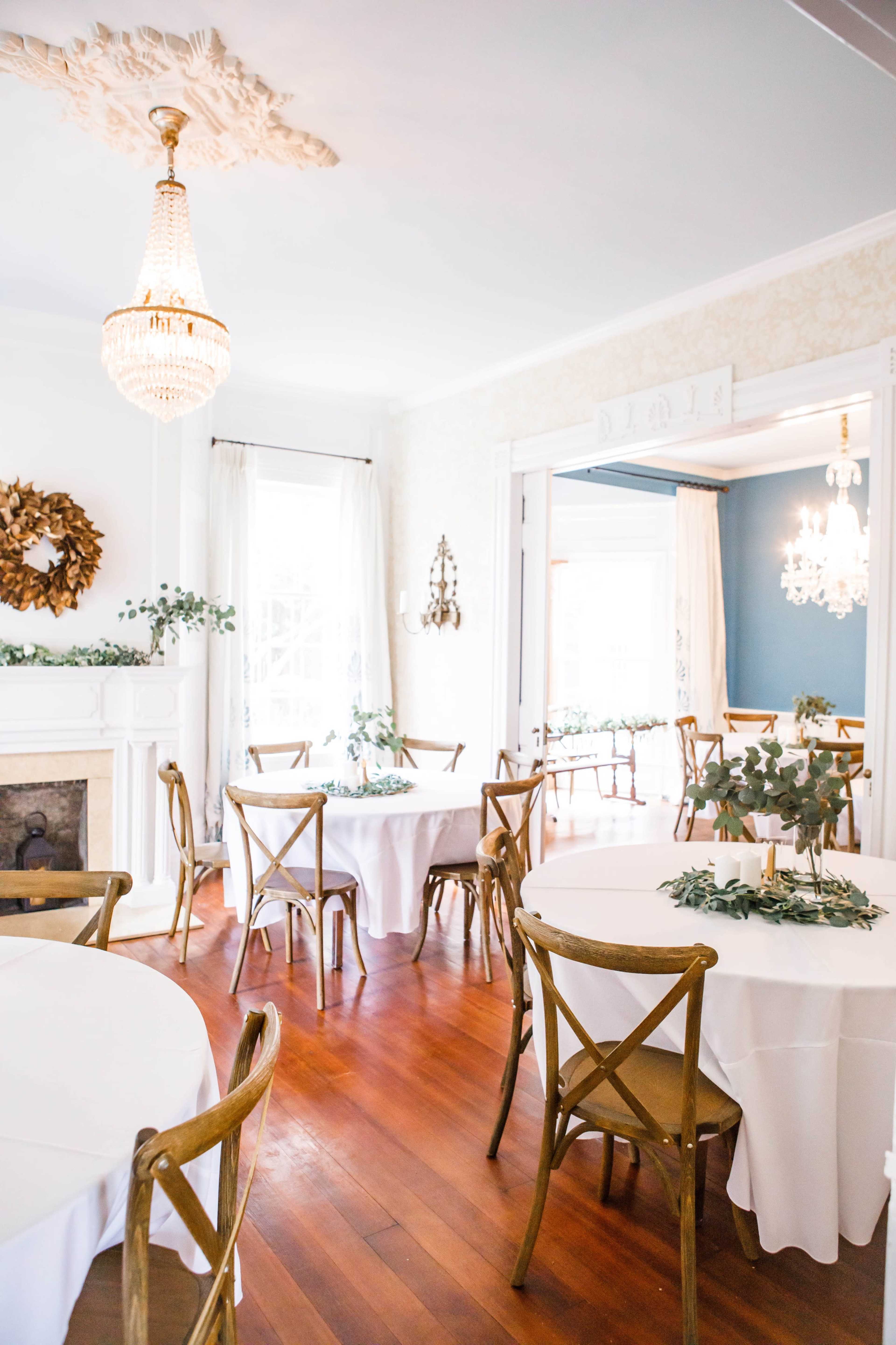 A bright dining room with wooden floors, round tables covered in white tablecloths, and decorative elements like a chandelier and wall decor.