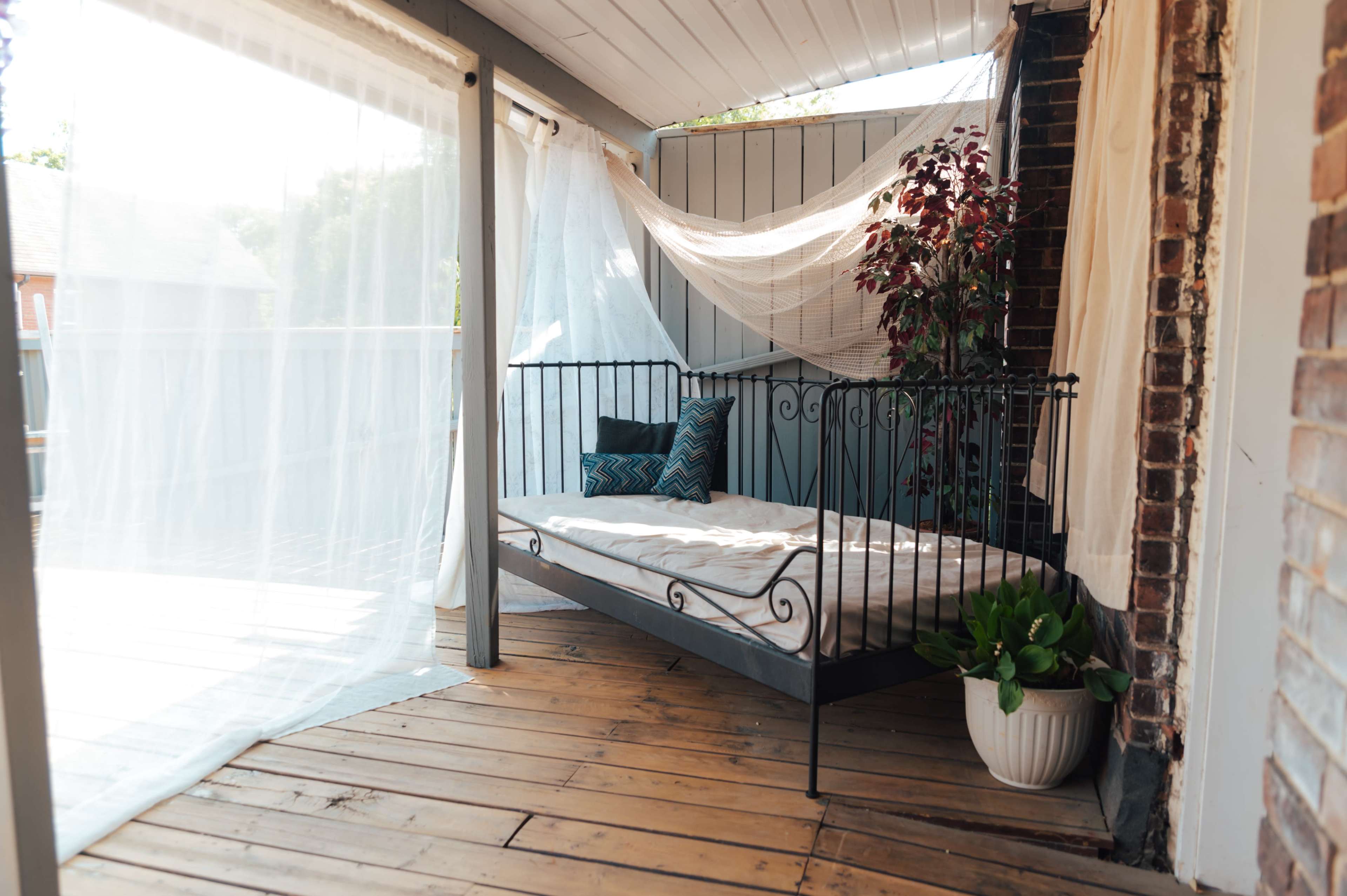The image shows a small, shaded outdoor area featuring a metal bed frame with a mattress, surrounded by sheer curtains and plants, on a wooden deck.