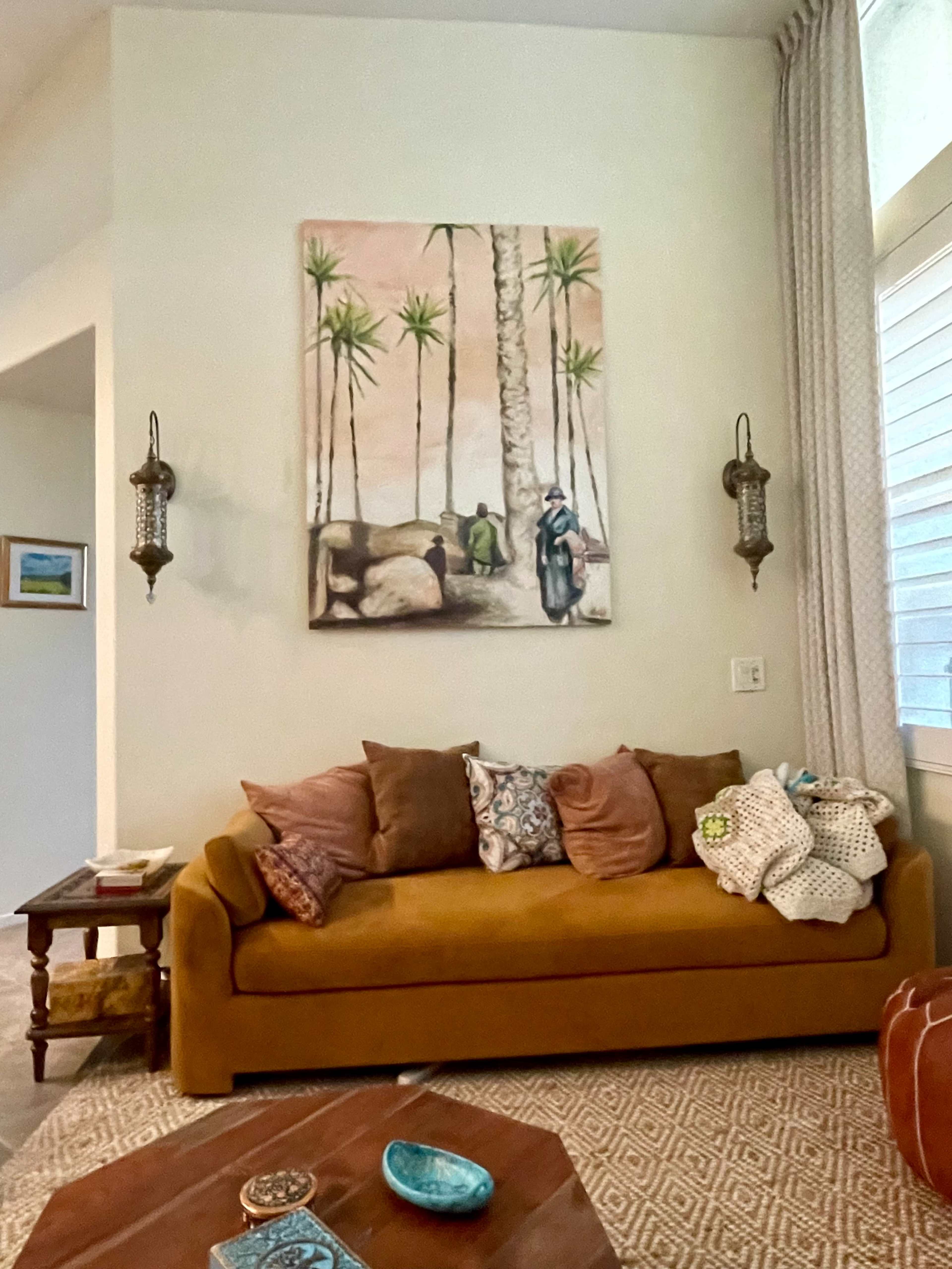 The image shows a living room featuring a mustard-colored sofa adorned with decorative pillows, a wooden coffee table, and a large palm tree artwork on the wall.