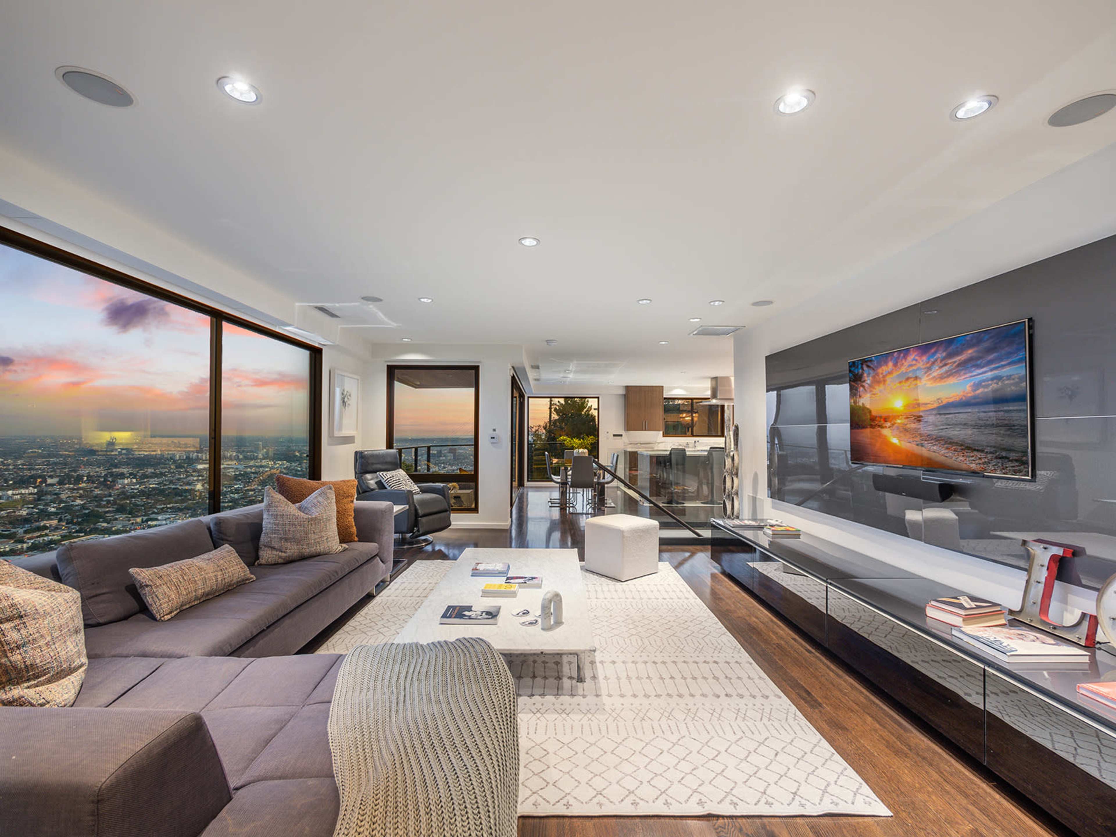 A modern living room features large windows with city views, a sectional sofa, a coffee table, and a wall-mounted television.