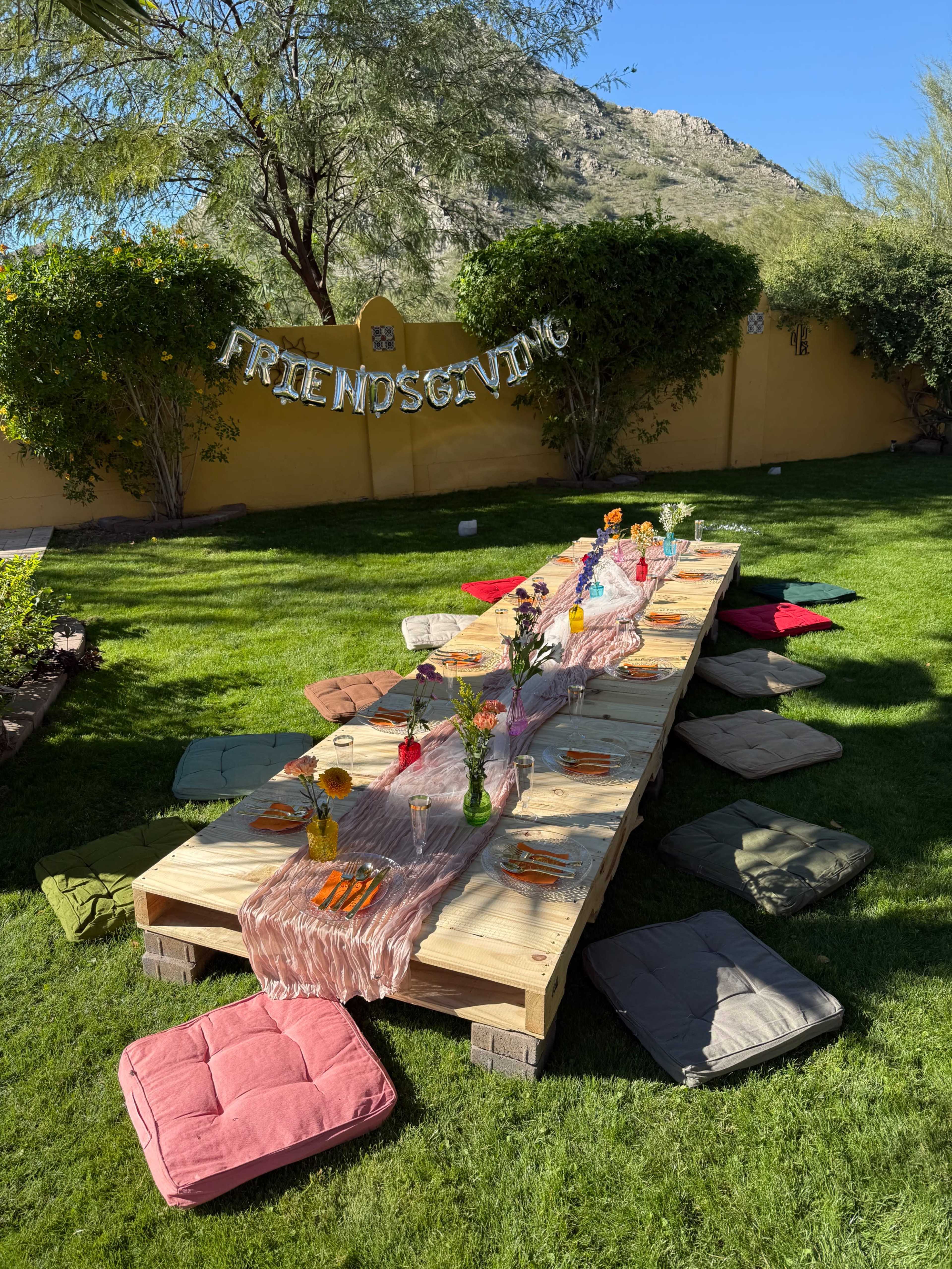 A long, rustic dining table is set outdoors on a grassy area, surrounded by colorful cushions, with decorative glasses and plates arranged along the table.
