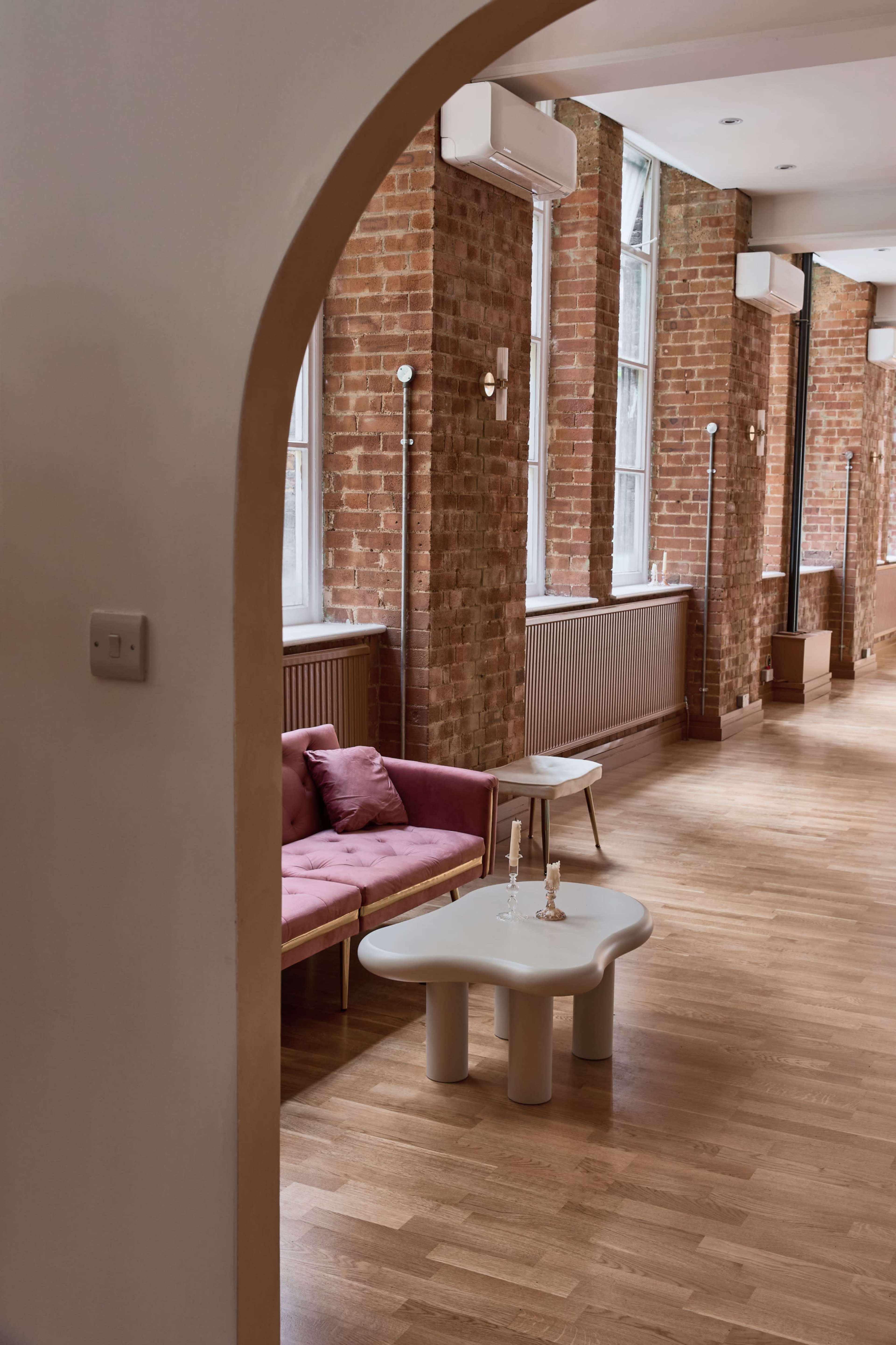The image shows a well-lit room with exposed brick walls, featuring a pink sofa, a round coffee table, and large windows along one side.
