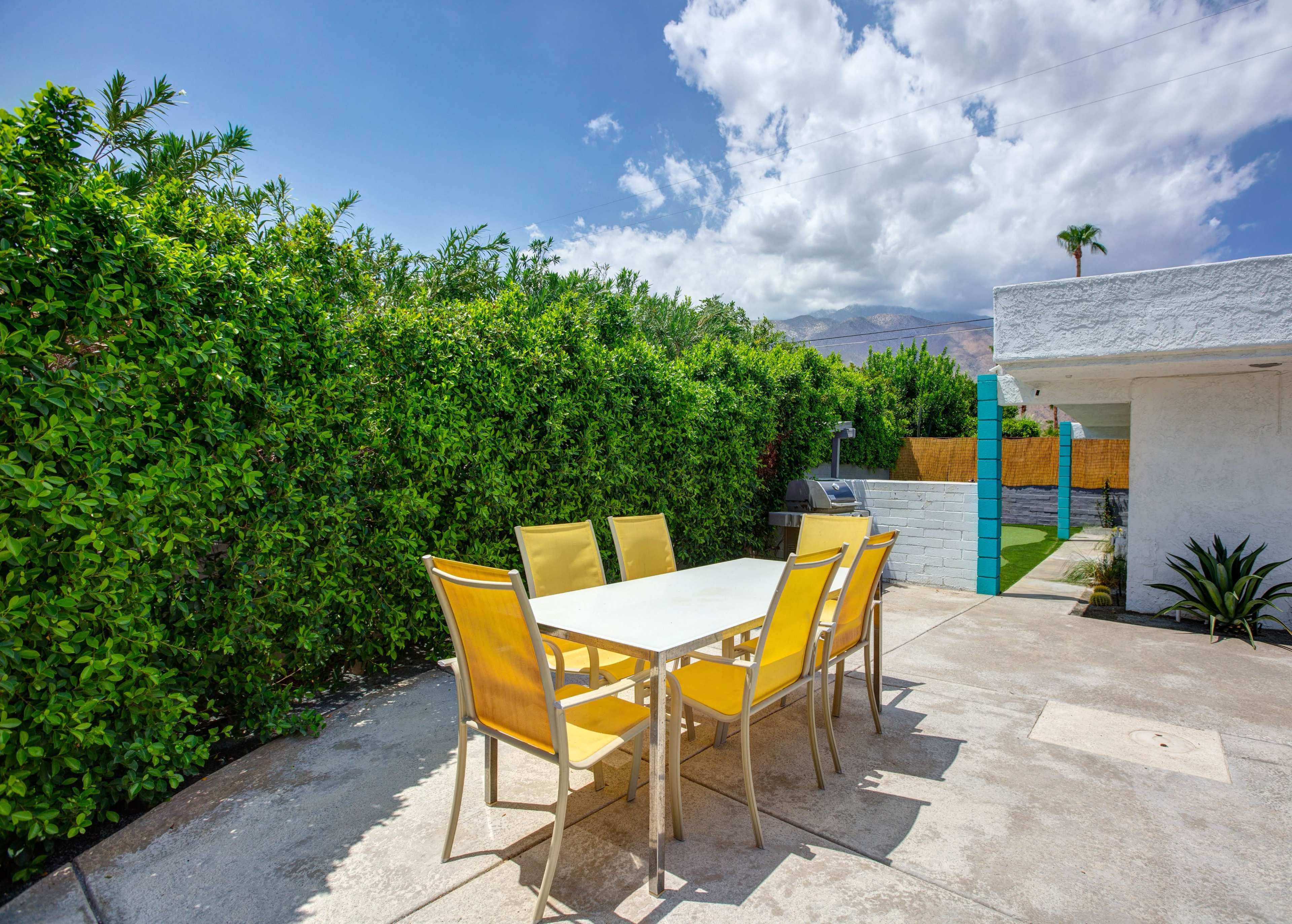 A patio area features a table surrounded by yellow chairs, bordered by lush green hedges and a sunny sky.