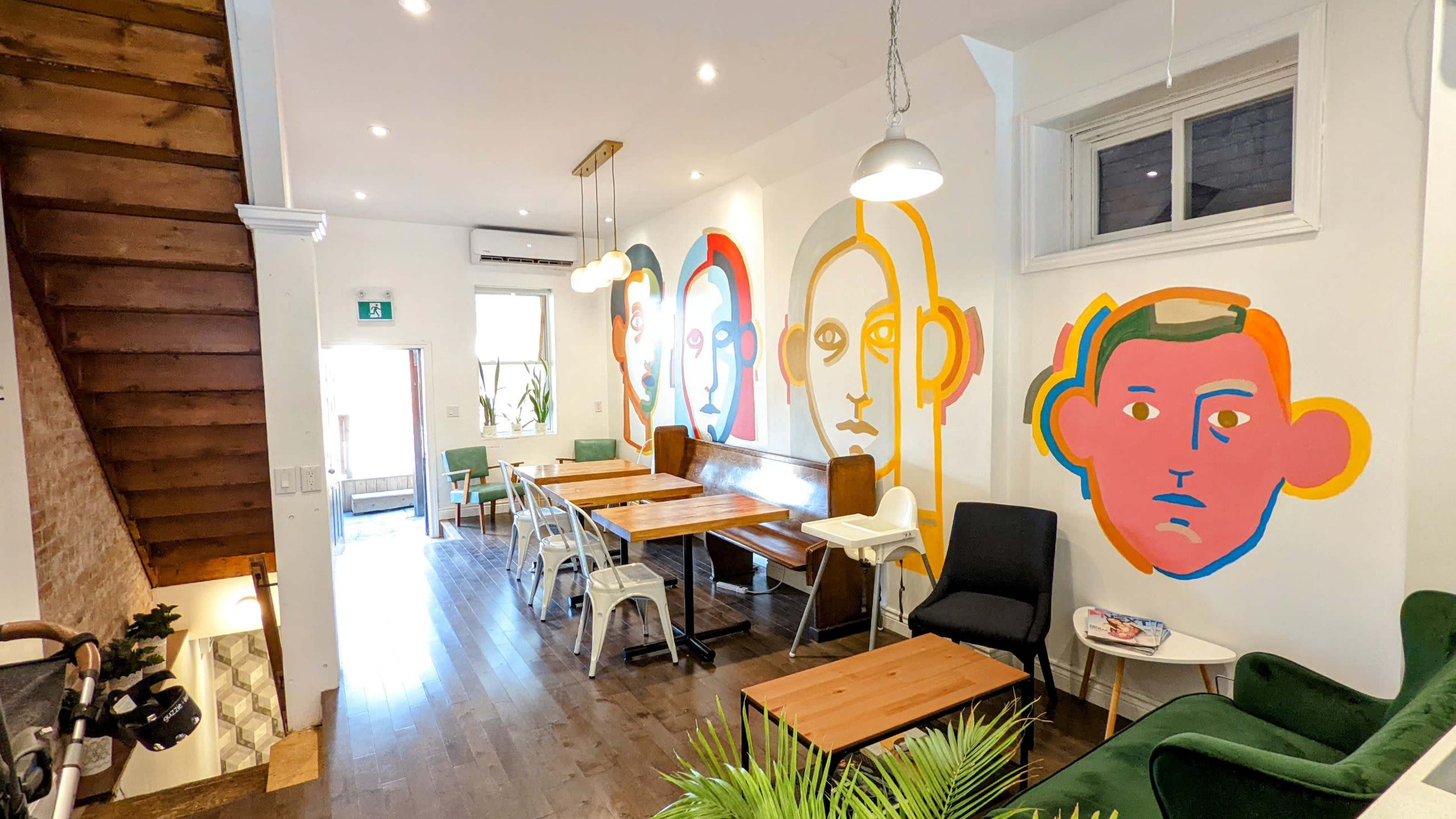A modern café interior featuring wooden tables, a staircase, and colorful mural artwork of stylized faces on the walls.