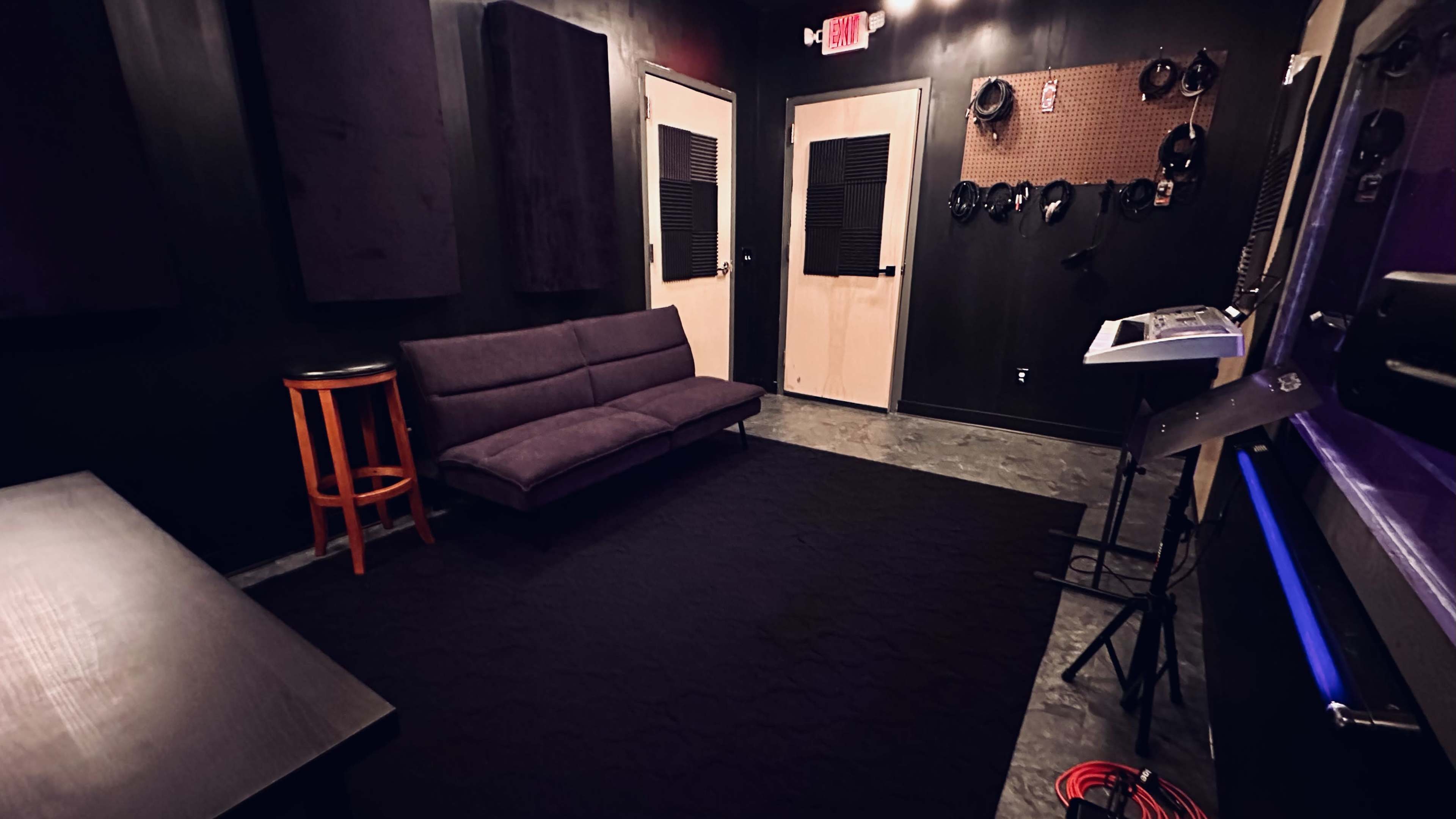 A small recording studio featuring black walls, a purple couch, soundproofing panels, and musical equipment arranged against the walls.
