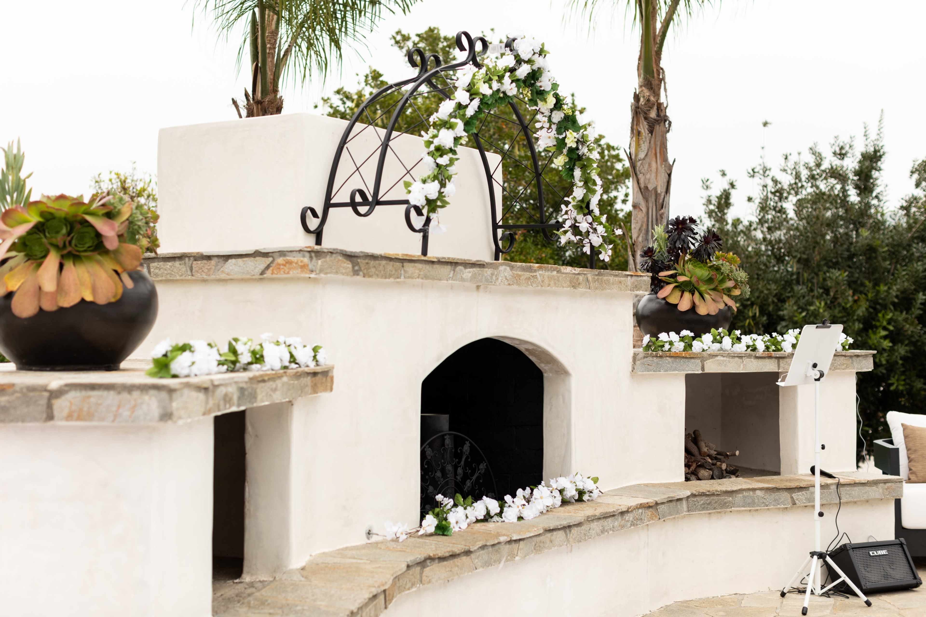 A stone structure with a decorative arch adorned with white flowers, surrounded by potted plants and a seating area.