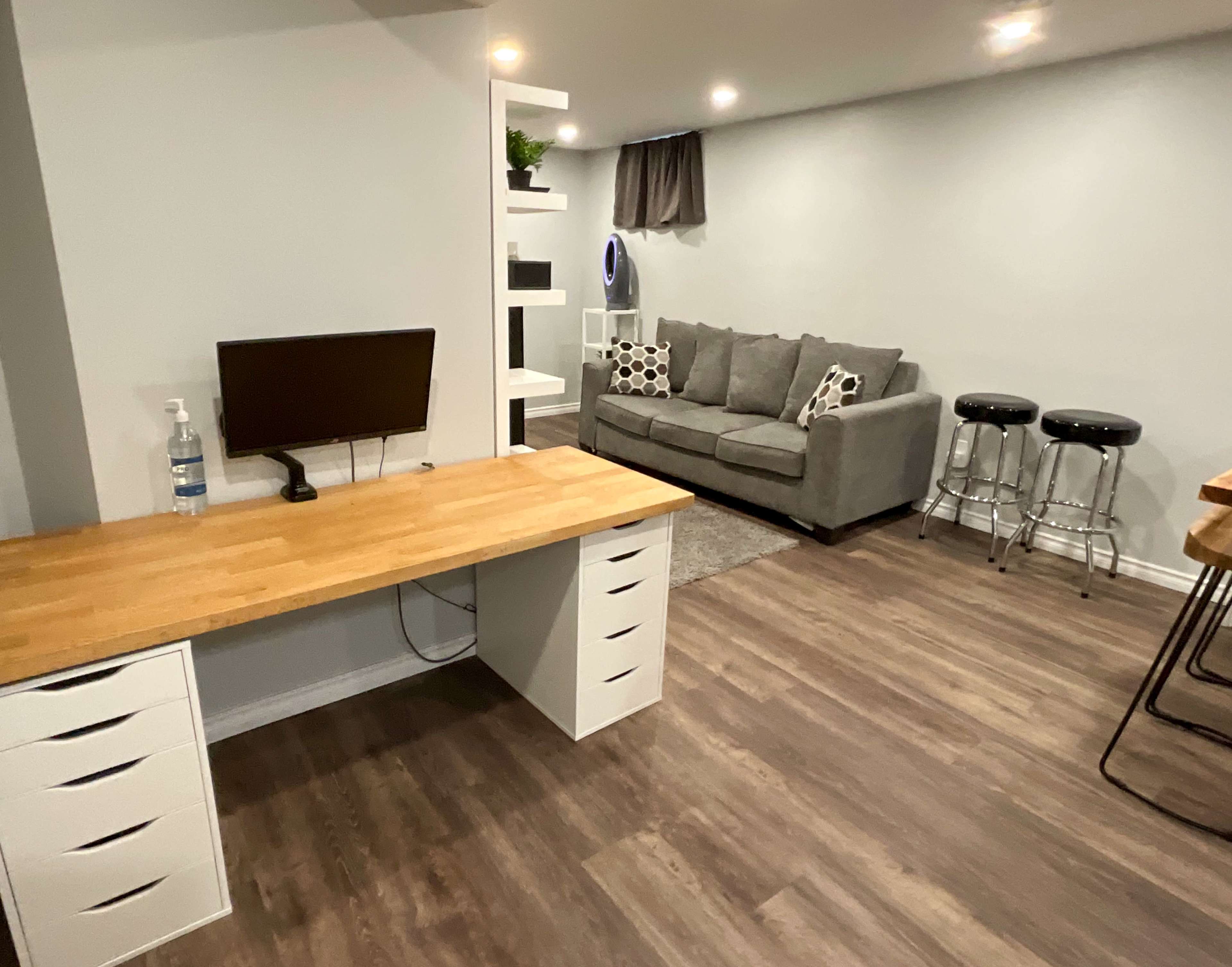 The image shows a modern interior space featuring a wooden desk with a computer monitor, a gray sofa, two bar stools, and a shelf against the wall.
