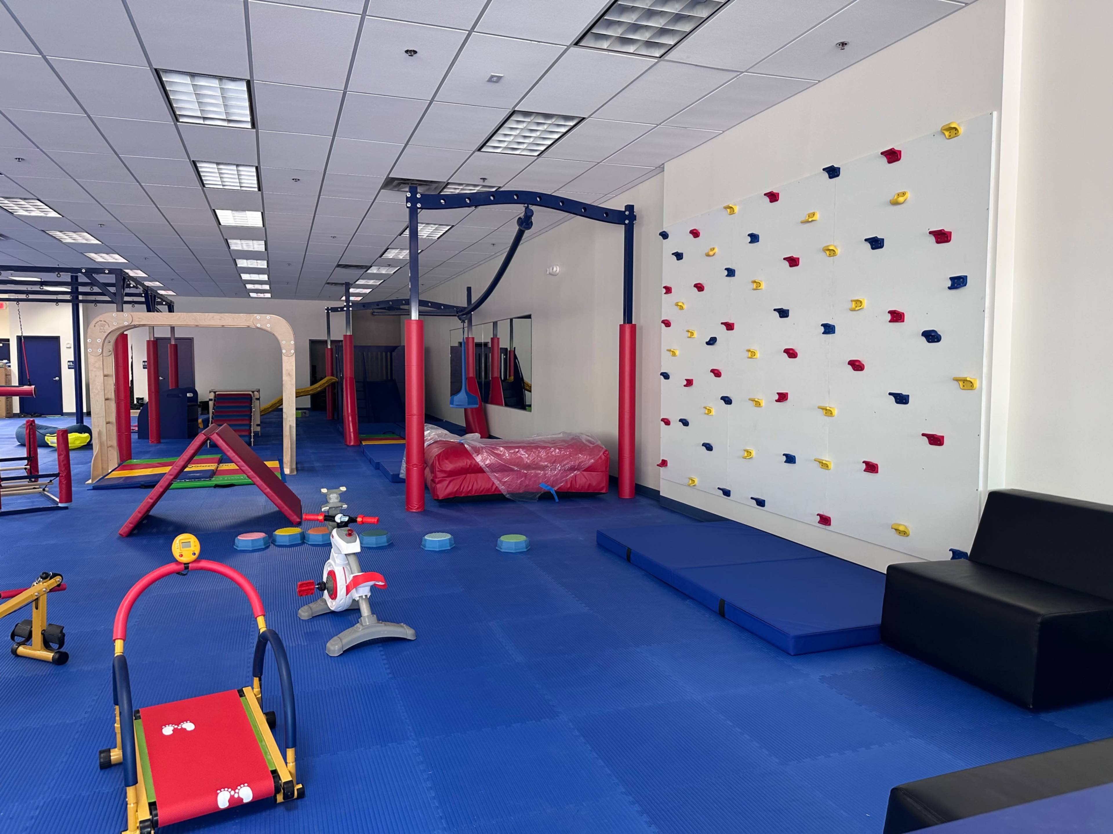 The image shows a brightly colored indoor play area featuring climbing structures, a small climbing wall, and various toys spread across blue mats.