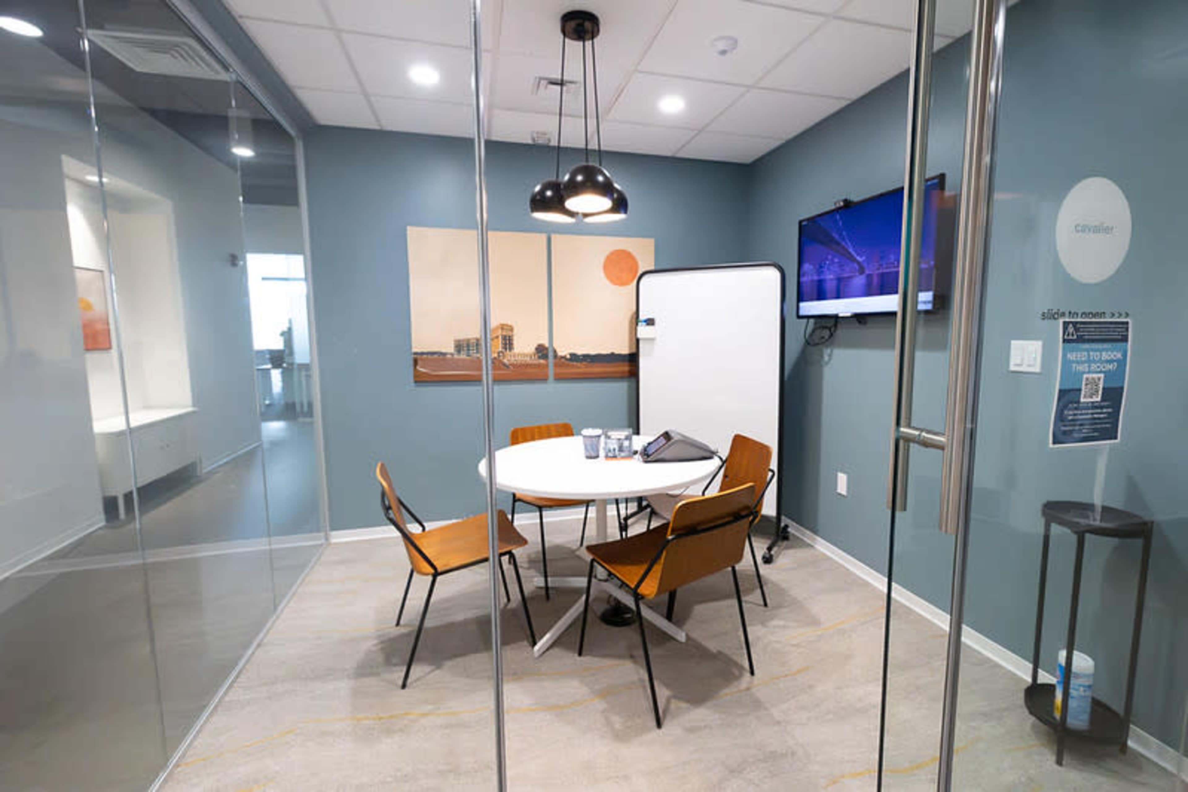 The image shows a small conference room with a round table, four chairs, a whiteboard, and a television mounted on the wall.