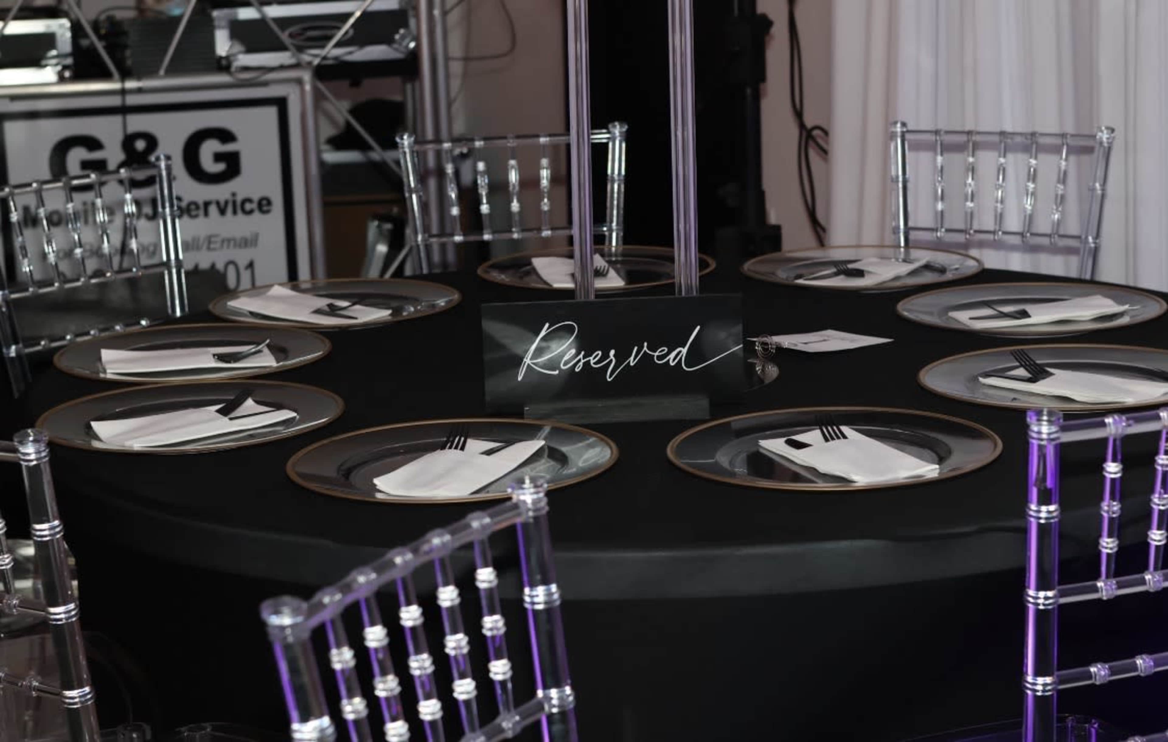 A round table covered with a black cloth features reserved seating indicated by a sign and elegant plates and utensils set for dining.