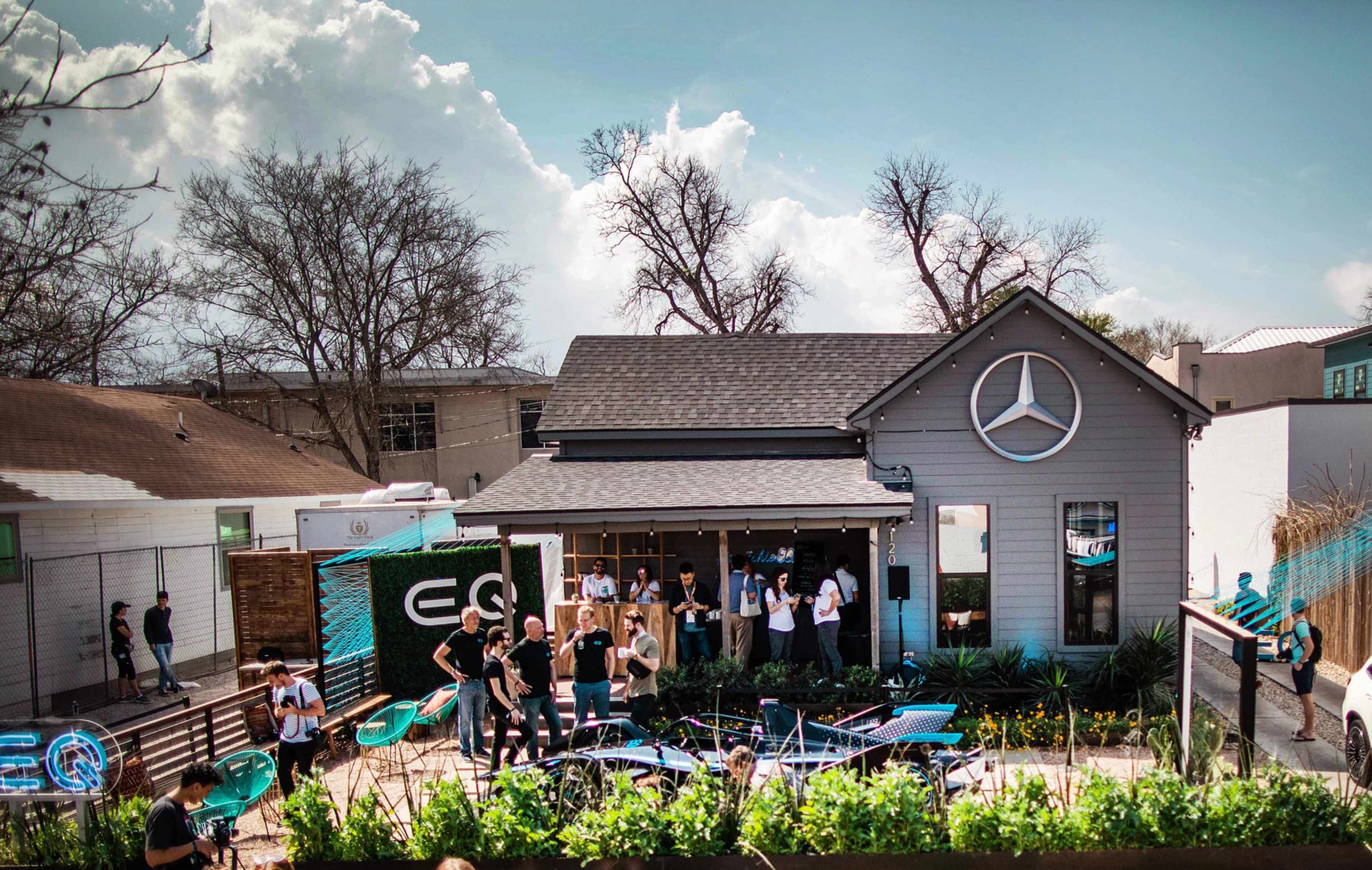 A group of people socializes outside a modernized house featuring a Mercedes-Benz logo, surrounded by greenery and patio furniture.