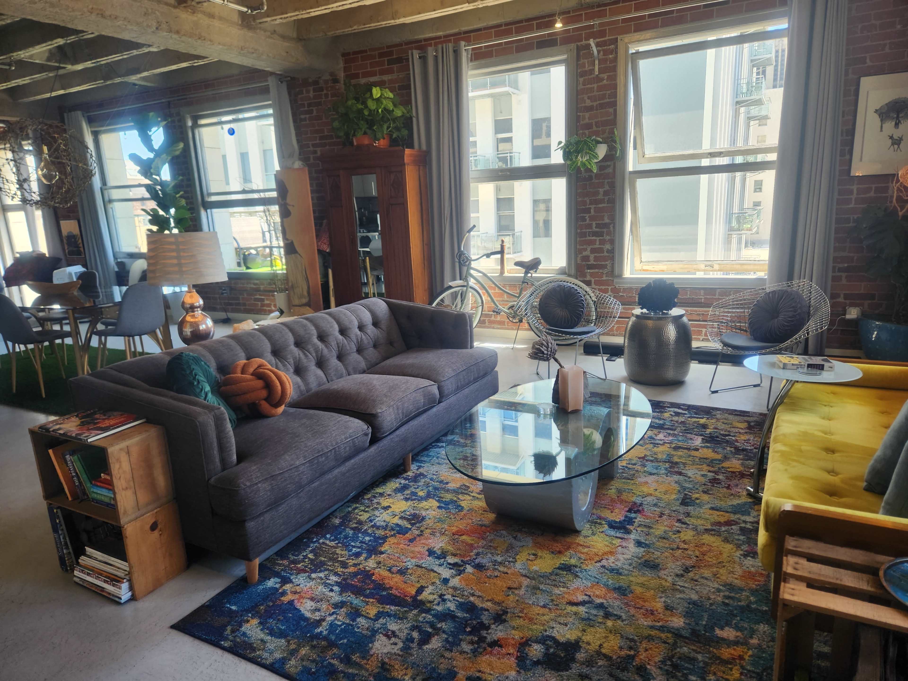 The image shows a spacious loft-style living area featuring a gray sofa, a glass coffee table, and large windows that allow natural light to fill the room, alongside plants and various furniture pieces.