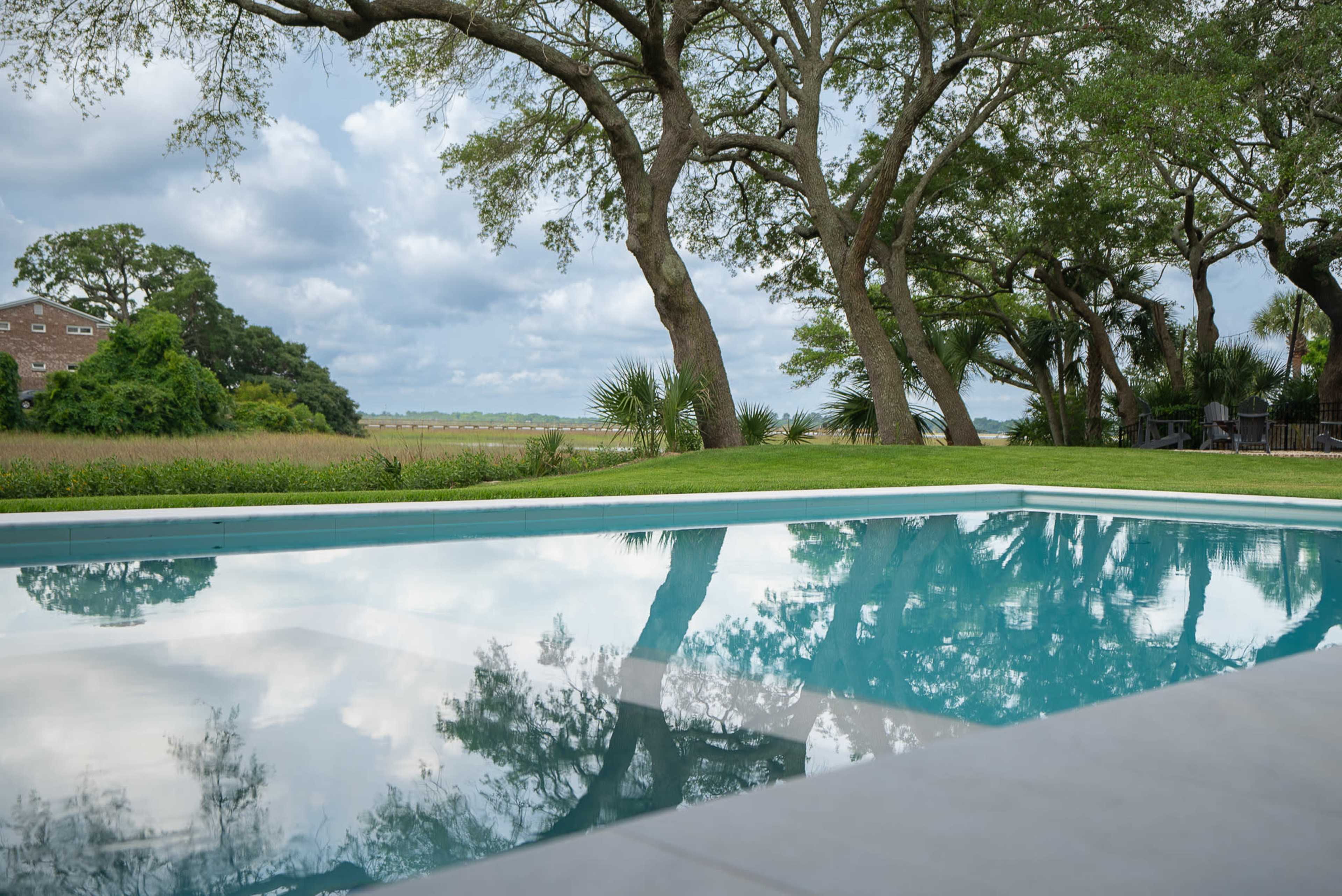 A swimming pool reflects the surrounding trees and cloudy sky in a green lawn area.