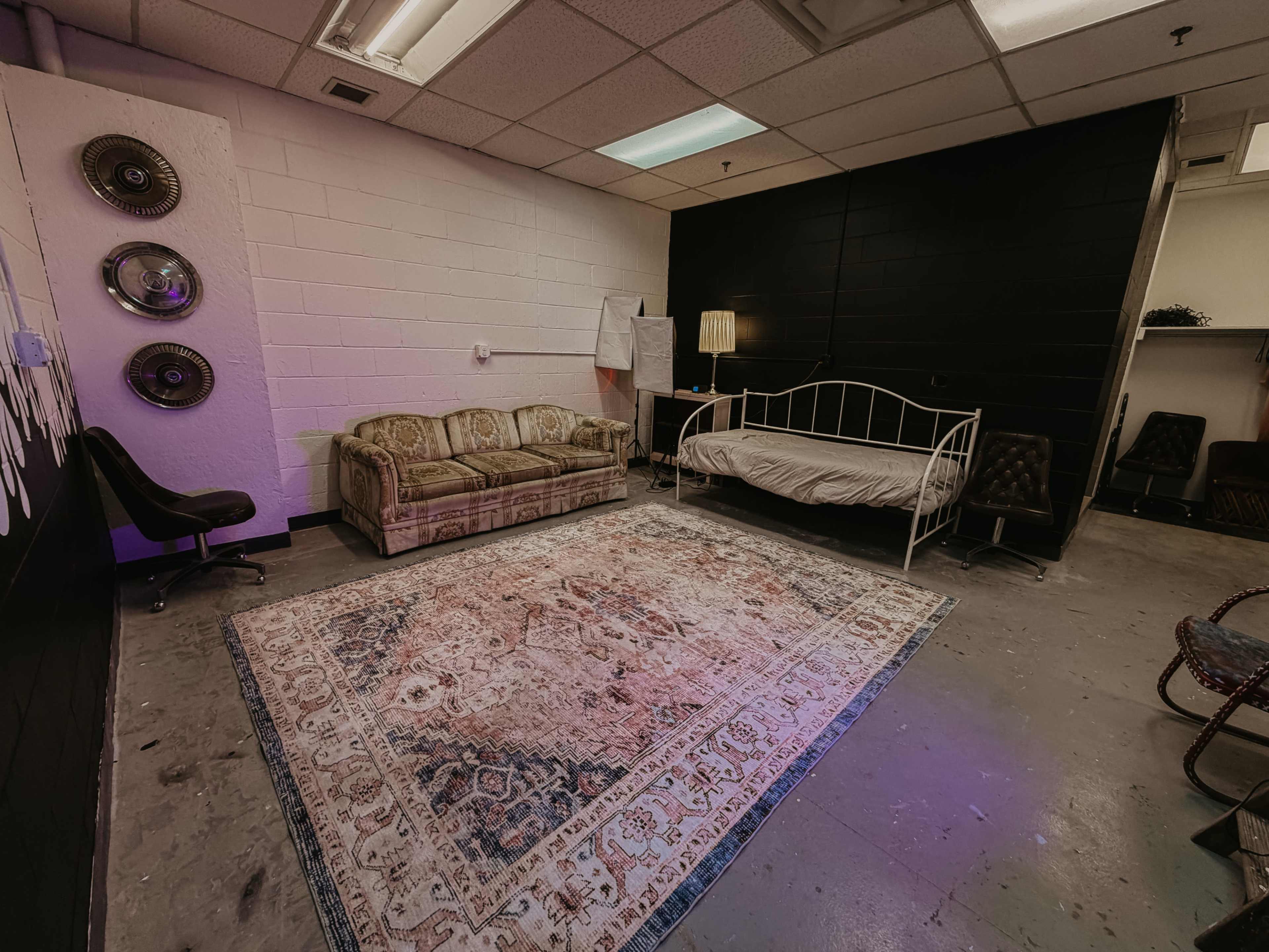 The image shows a sparse room containing a vintage couch, a bed with white bedding, and a large area rug on the concrete floor.