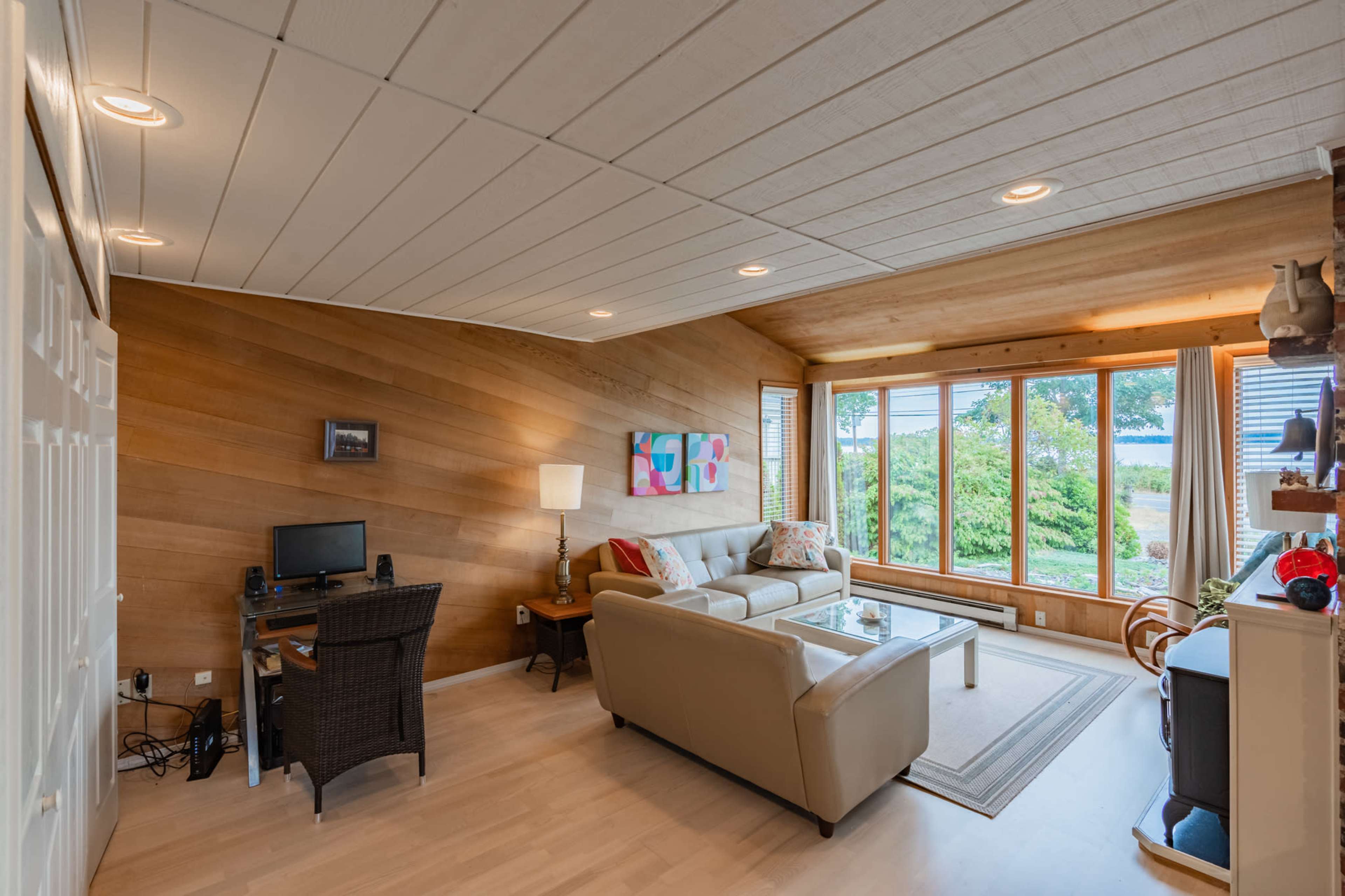 The image shows a living room with a beige sofa, a small desk with a computer, and large windows overlooking a garden.